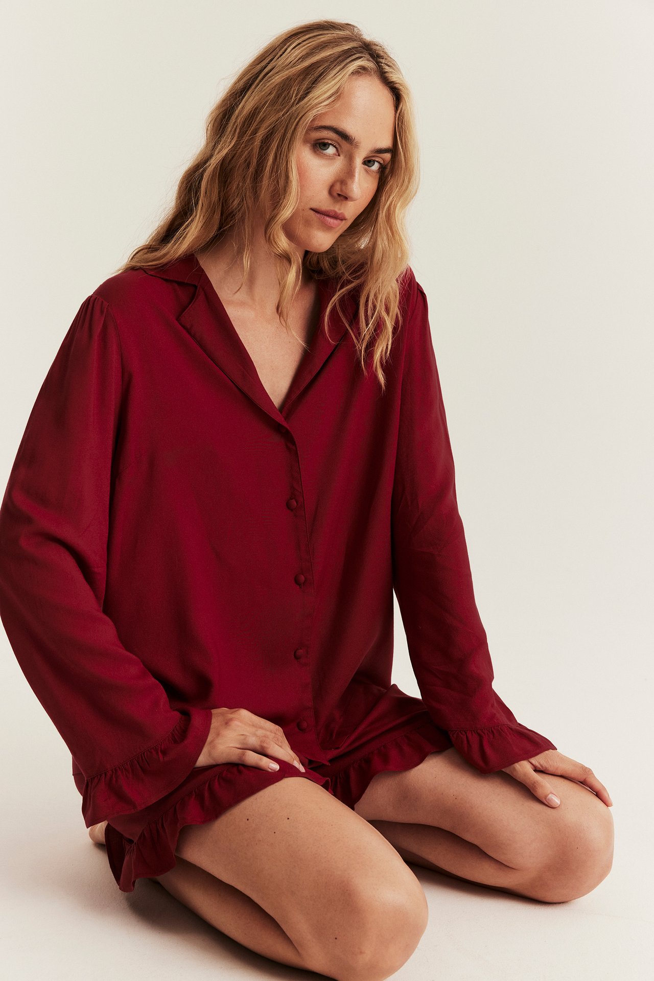 Frontansicht kniender Frau in rotem Viskose-Pyjama-Set mit Hemd und Rüschen-Shorts.