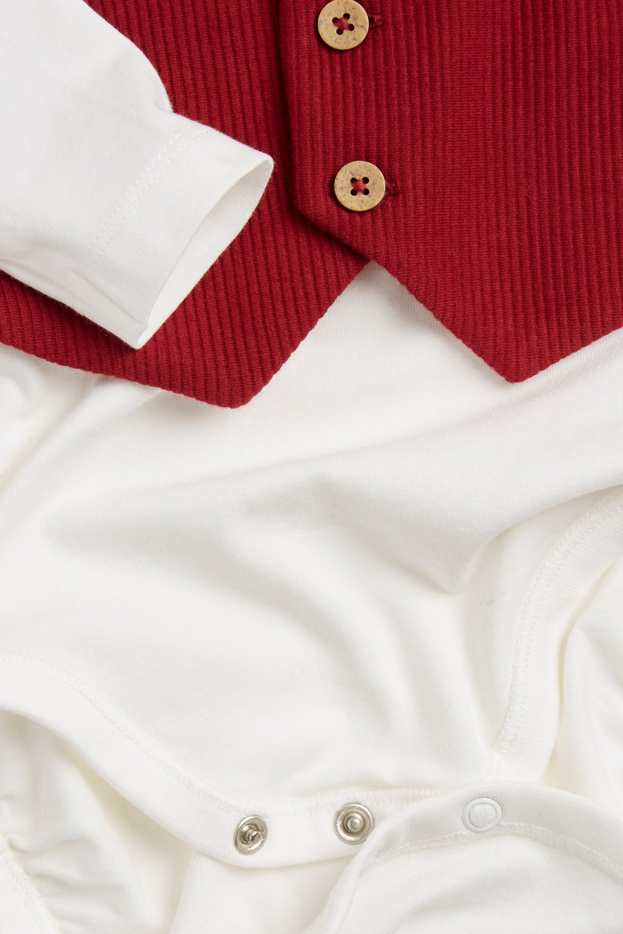 Close-up: baby's white long-sleeved cotton bodysuit with attached red ribbed vest and wooden buttons.
