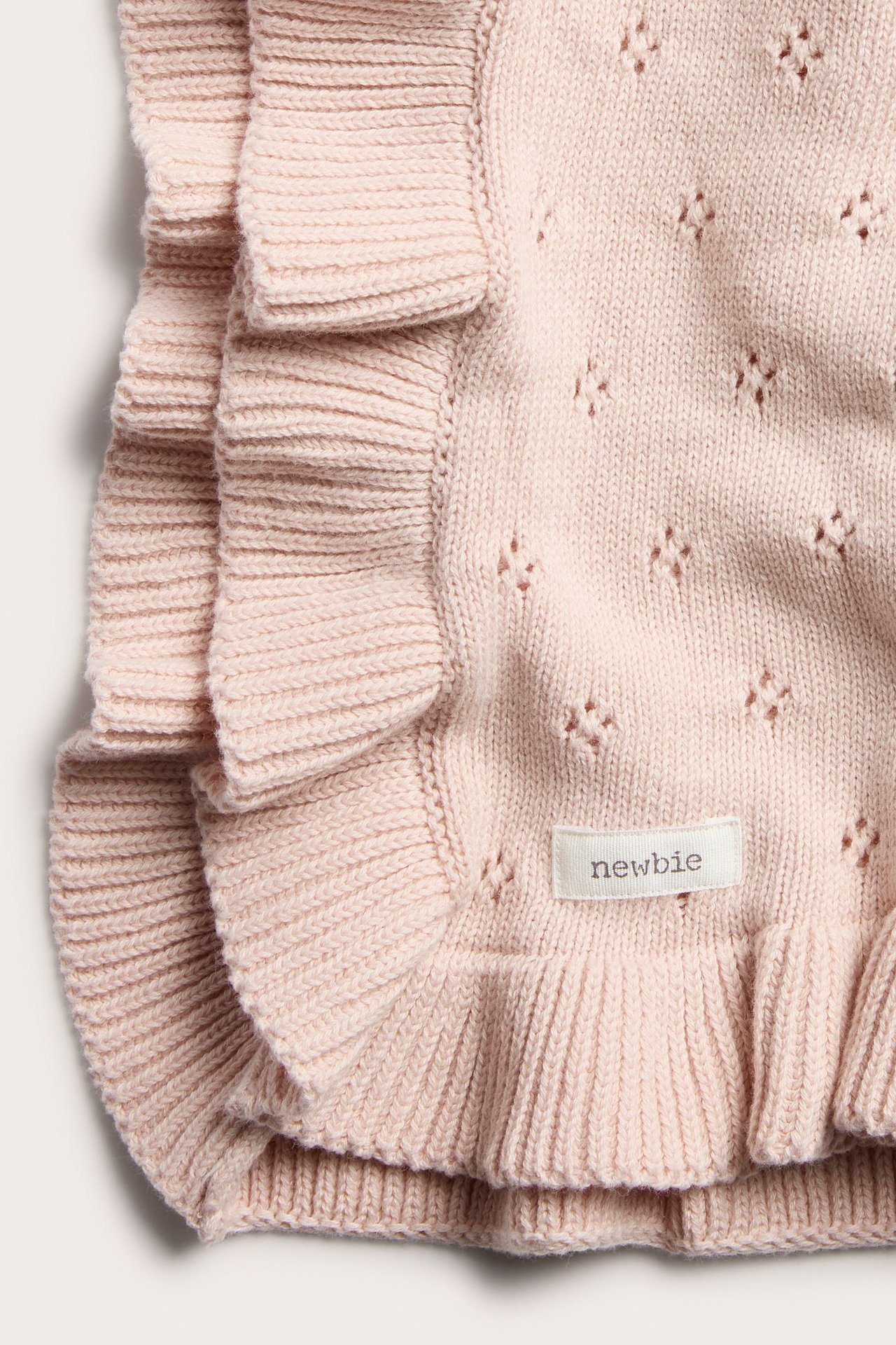 Nahaufnahme einer hellrosa gestrickten Baumwolldecke mit Lochmuster und Rüschenkanten, mit Newbie-Etikett.