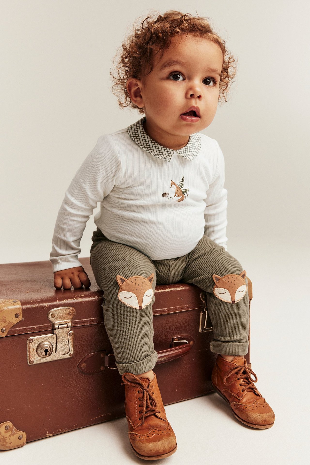 Baby in cremeweißem Ripp-Body, Fuchsdruck, kariertem Kragen. Olivgrüne Leggings mit Fuchs-Patches, braune Stiefel.