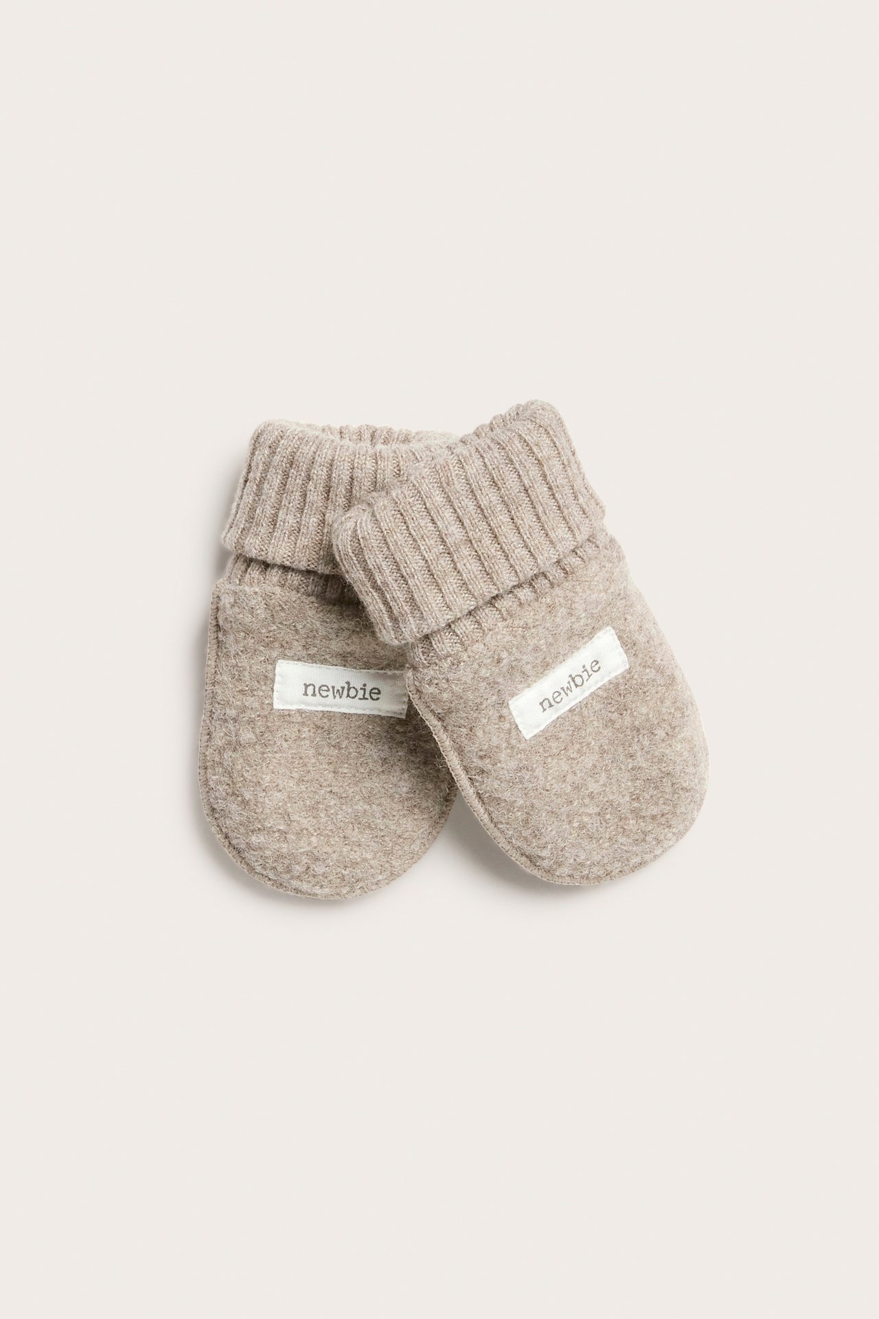 Pair of light brown baby mittens with ribbed cuffs and 'newbie' tags, top view.