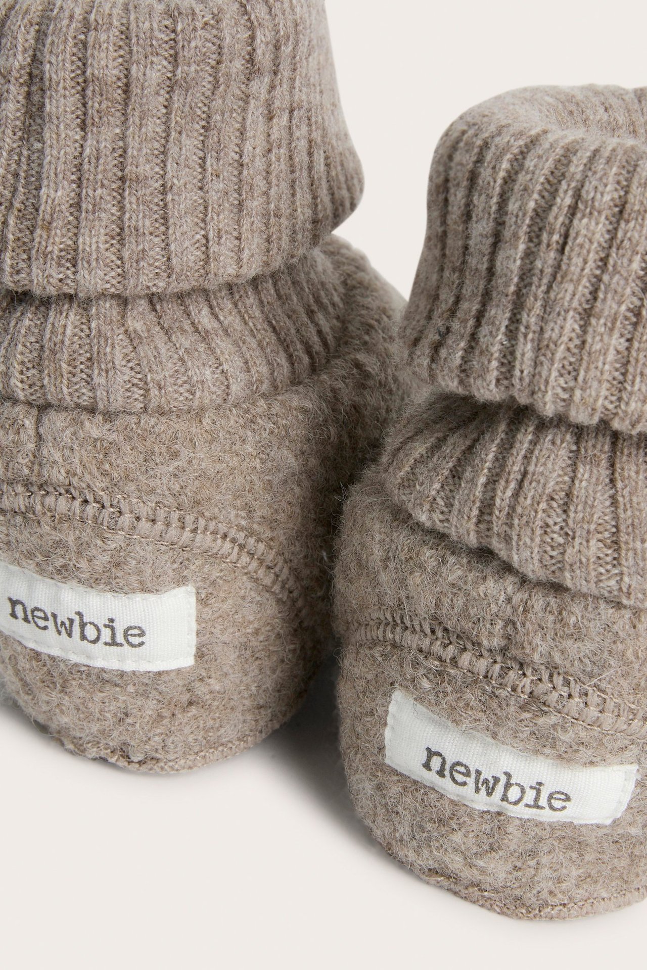 Close-up of two melange brown baby wool booties with ribbed, folded cuffs and white 'newbie' labels.