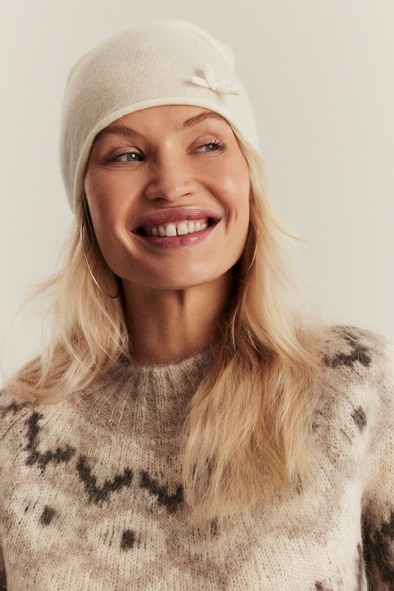 Front view: Woman in offwhite wool blend beanie with a bow, patterned knit sweater, and hoop earrings.