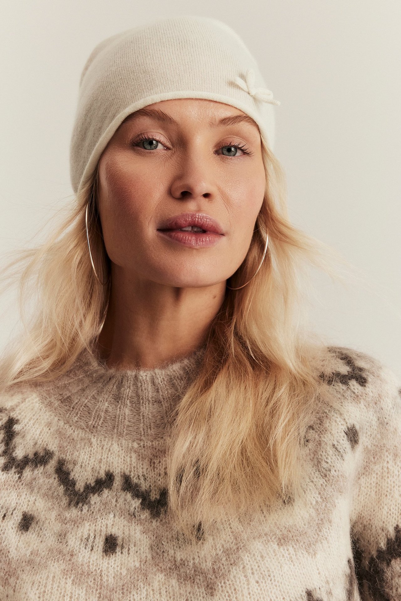 Close-up of woman in off-white wool beanie with bow, patterned sweater, and hoop earrings.