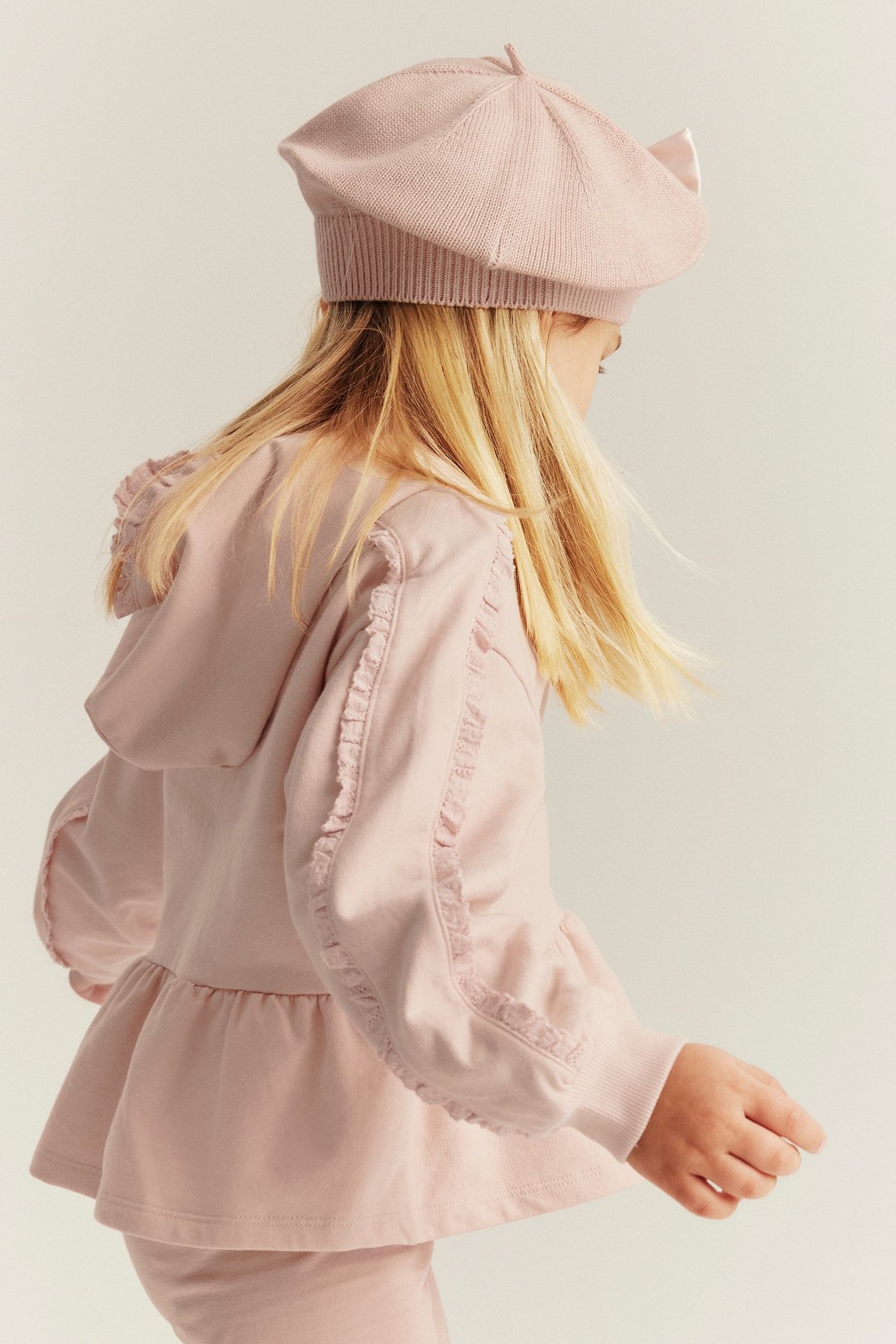 Side view of a kid in a light pink cotton hooded sweatshirt with lace trim and matching beret.