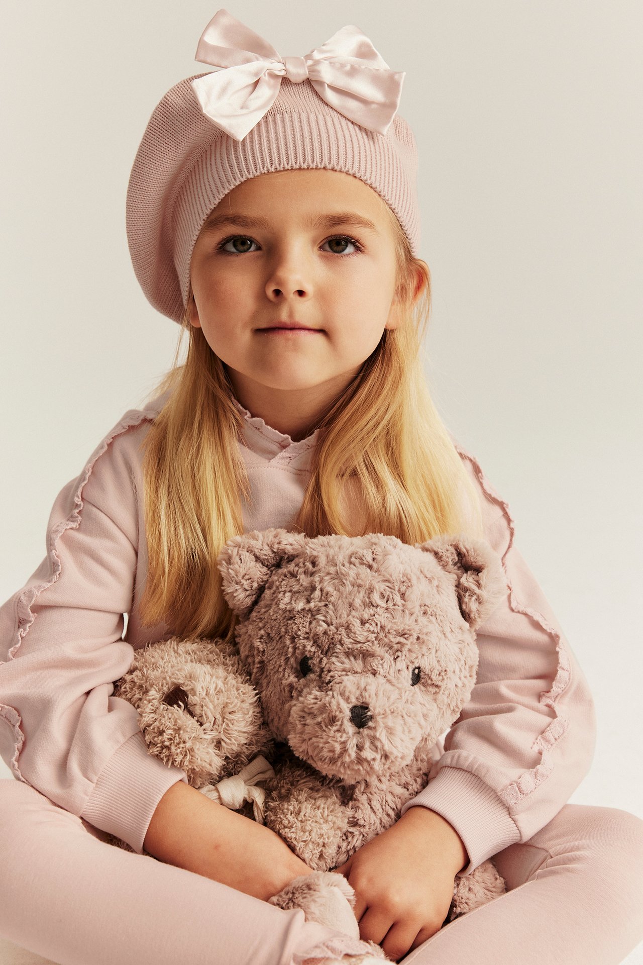 Vorderansicht: Mädchen in hellrosa Kapuzenpullover mit Spitze und passendem Barett, hält einen Teddybär.