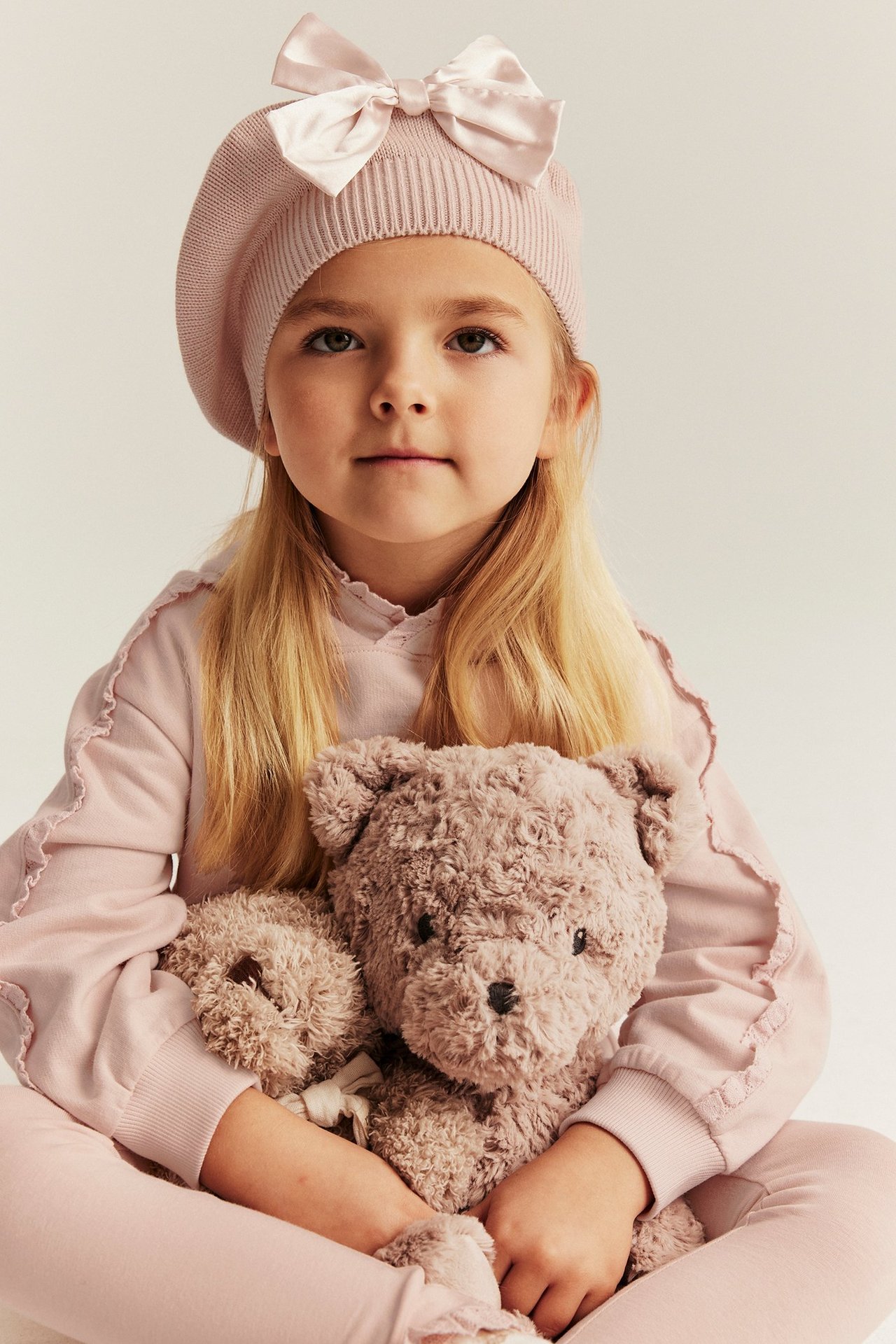 Vorderansicht: Mädchen in hellrosa Kapuzenpullover mit Spitze und passendem Barett, hält einen Teddybär.