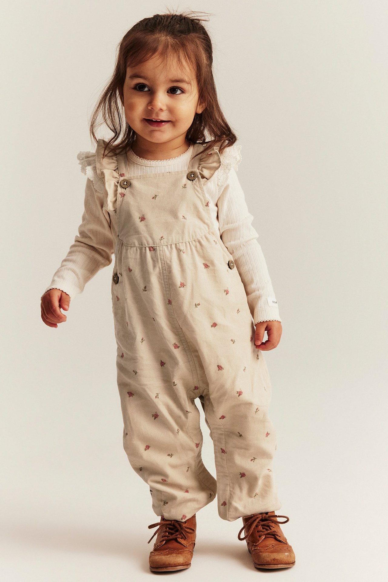 Front view of a kid in beige floral corduroy dungarees with ruffles, white long-sleeved top, and brown shoes.