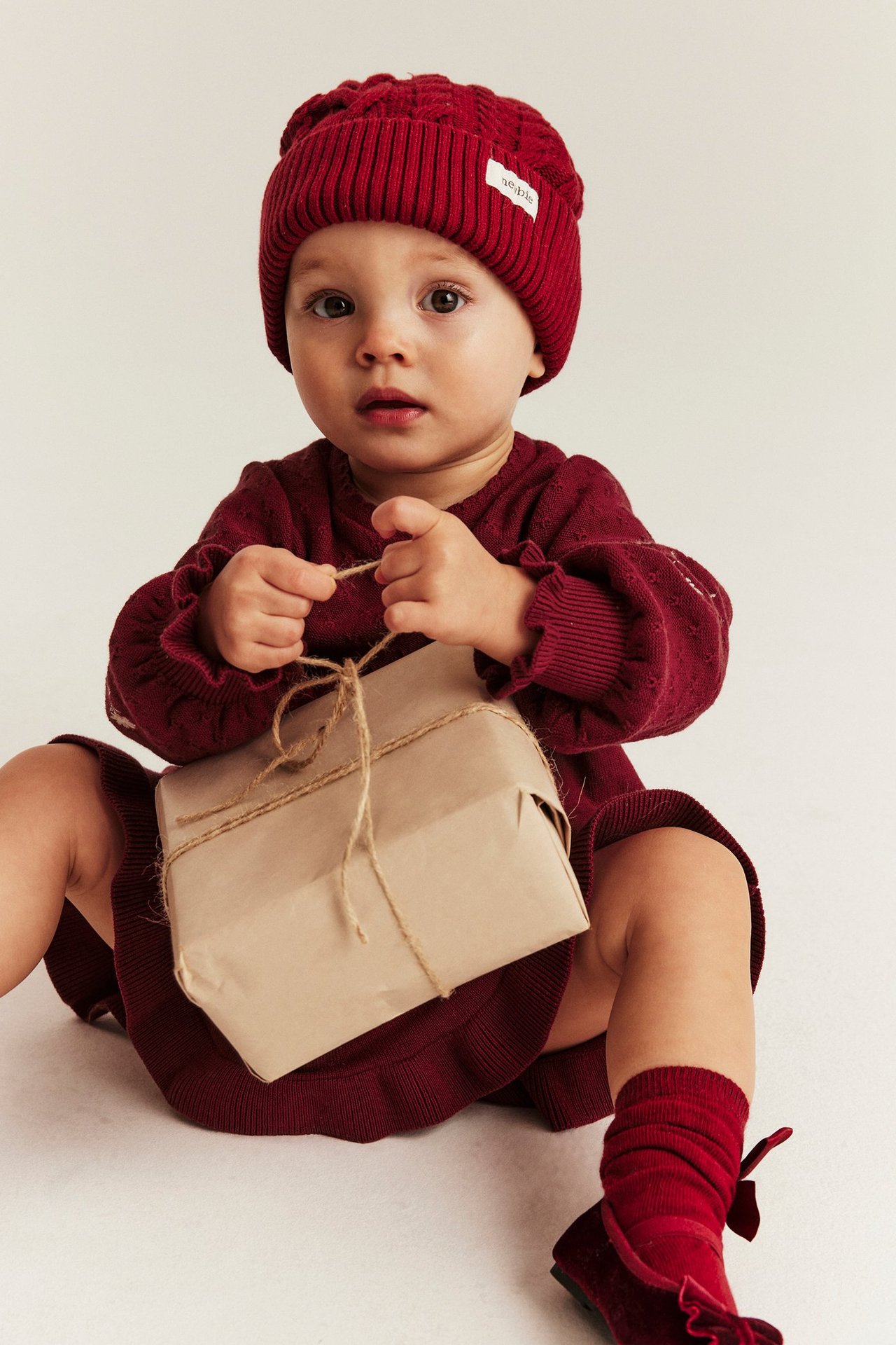 Baby boy in red knitted hat and sweater, holding a wrapped gift box.