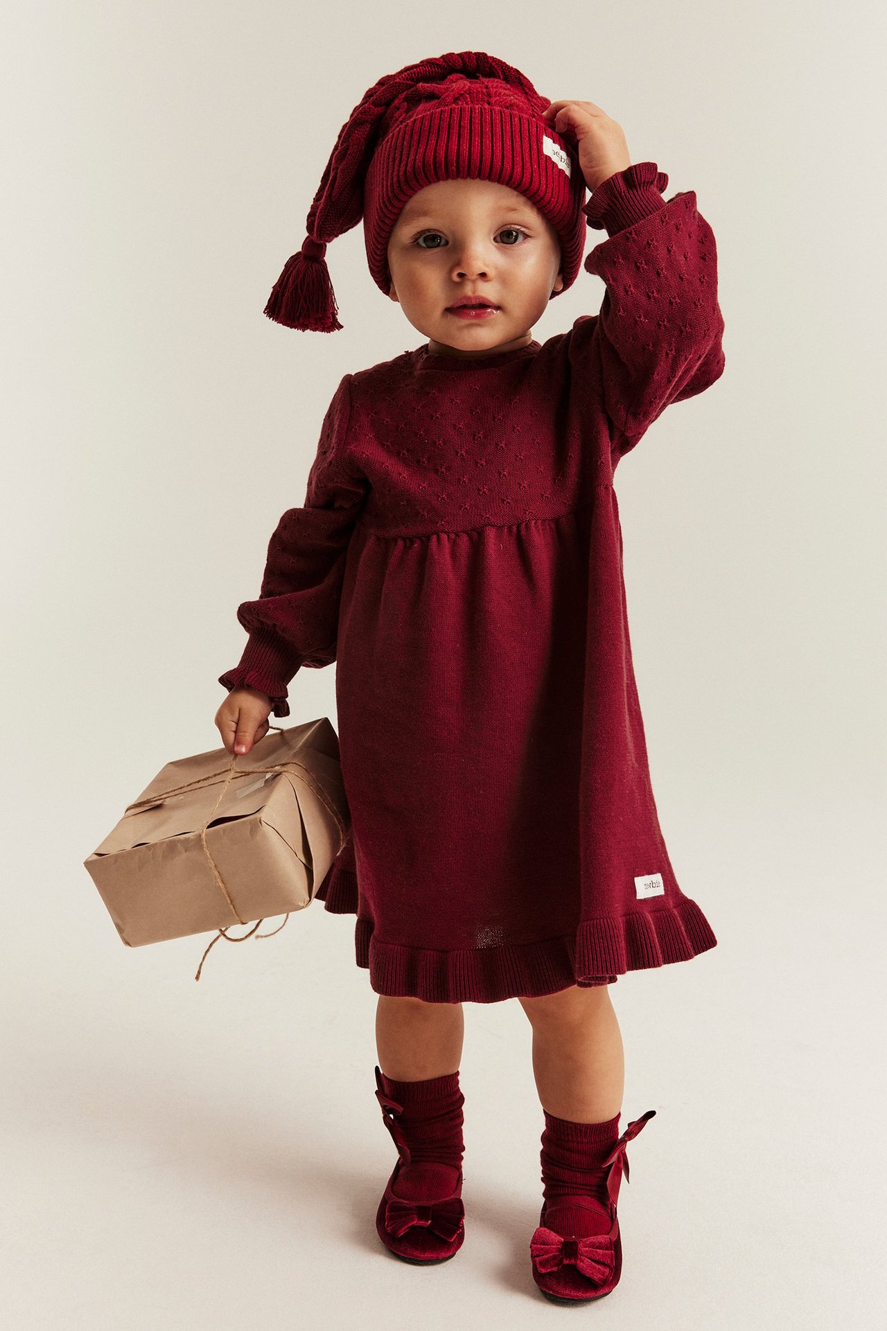 Young girl wearing a red knitted dress with embroidery on sleeves, ruffle hem, back buttons, and red socks and hat.