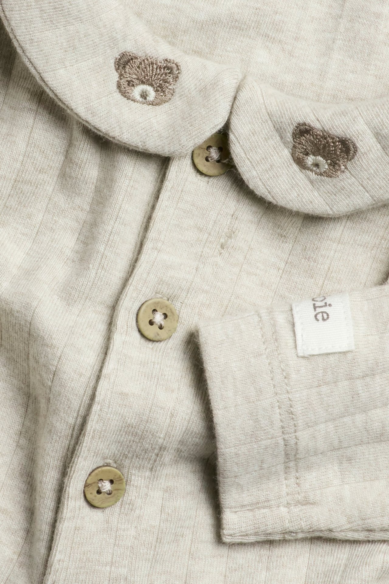 Nahaufnahme eines beigen gerippten Baby-Bodys mit Kragen, Teddybär-Stickerei und Knöpfen vorne.