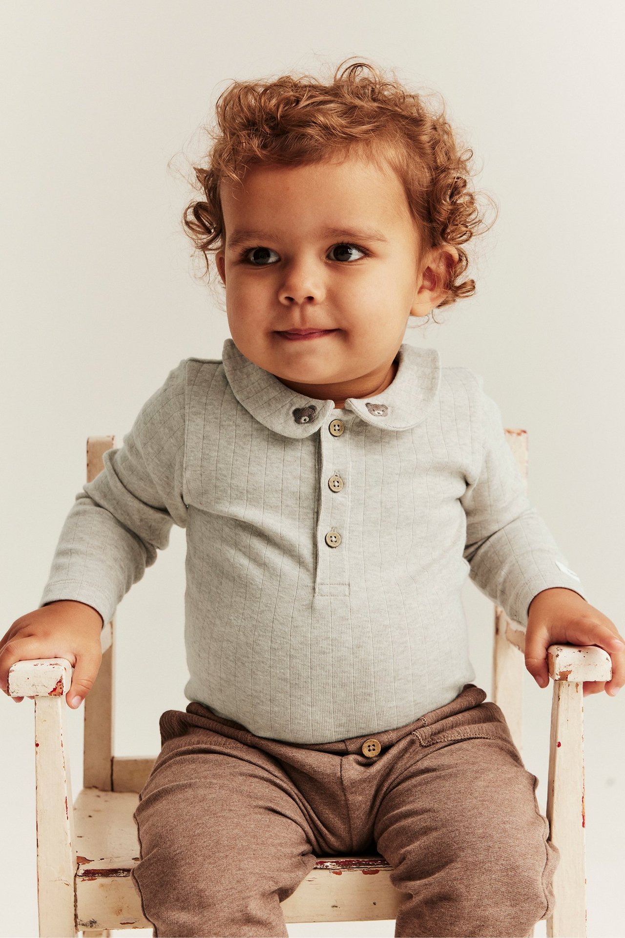 Front view of a baby wearing a light grey ribbed bodysuit with teddy bear embroidered collar and brown trousers.