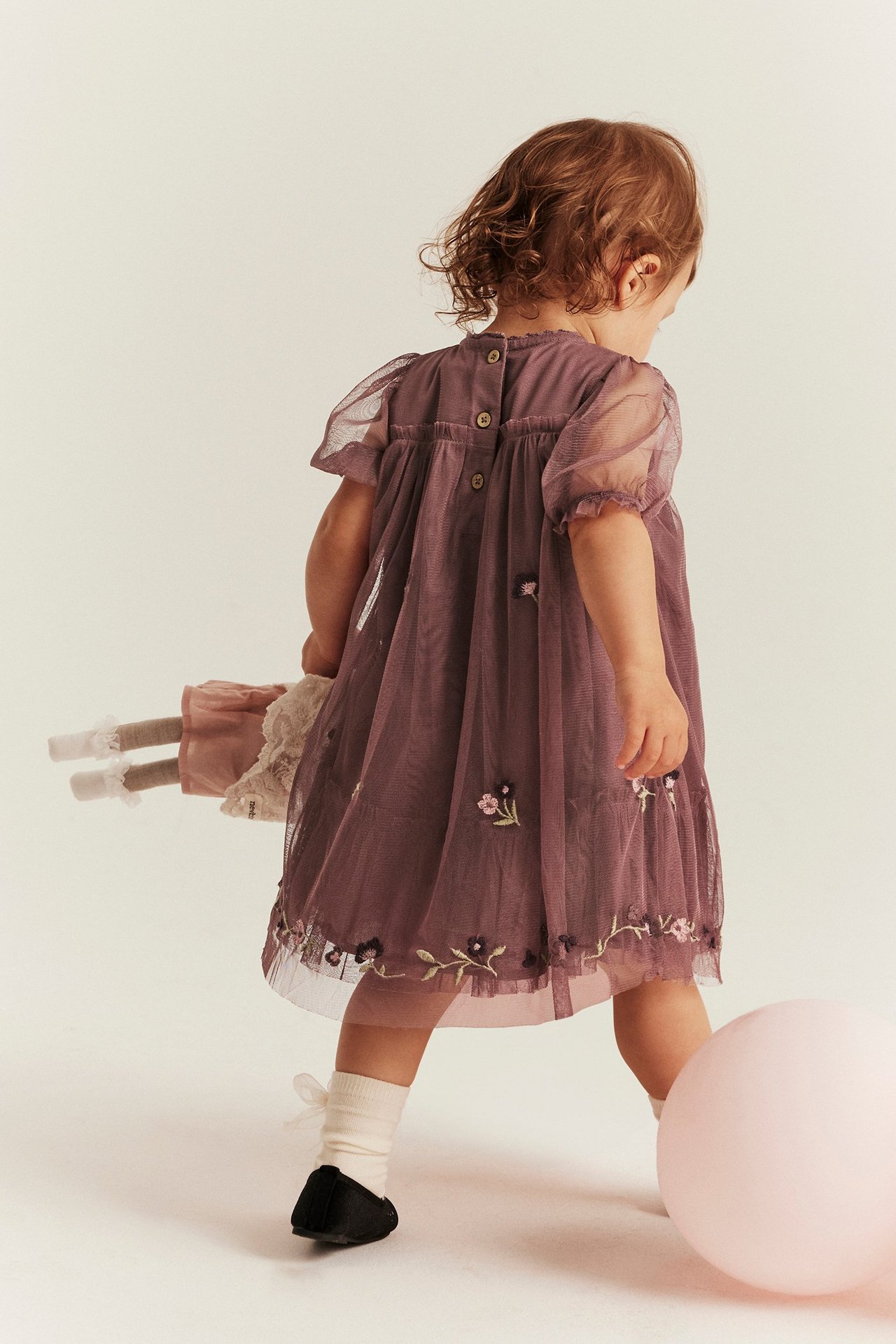 Rückenansicht: Baby im dunkelrosa bestickten Netz-Kleid mit Puffärmeln, weißen Socken, schwarzen Schuhen, hält Puppe.