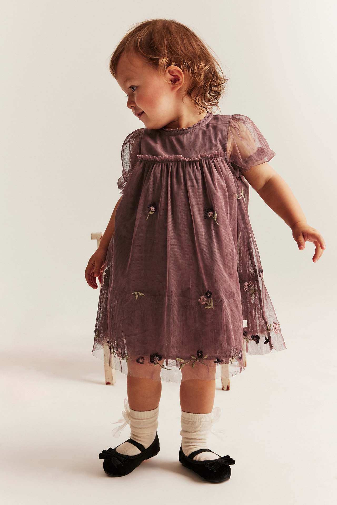Front view of a baby wearing a dark pink mesh dress with floral embroidery, puff sleeves, cream socks, and black ballet shoes.