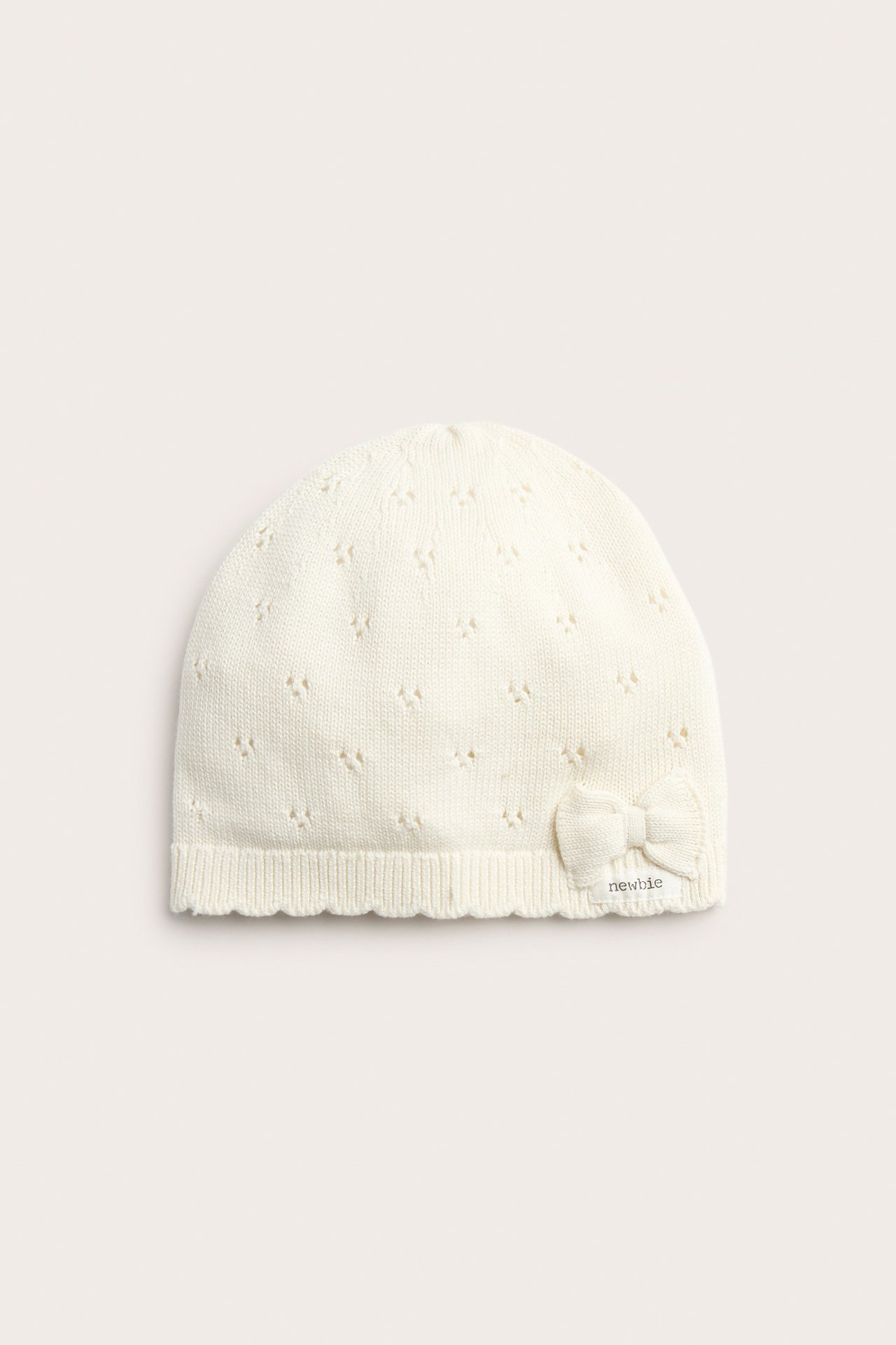 Vorderansicht, cremefarbene gemusterte Strickmütze für Baby/Kind mit Schleife und Wellenrand.