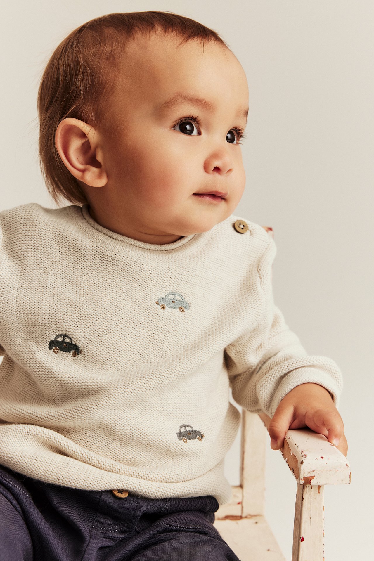 Close-up front view of a baby in a beige knitted sweater with car embroidery and dark blue pants.