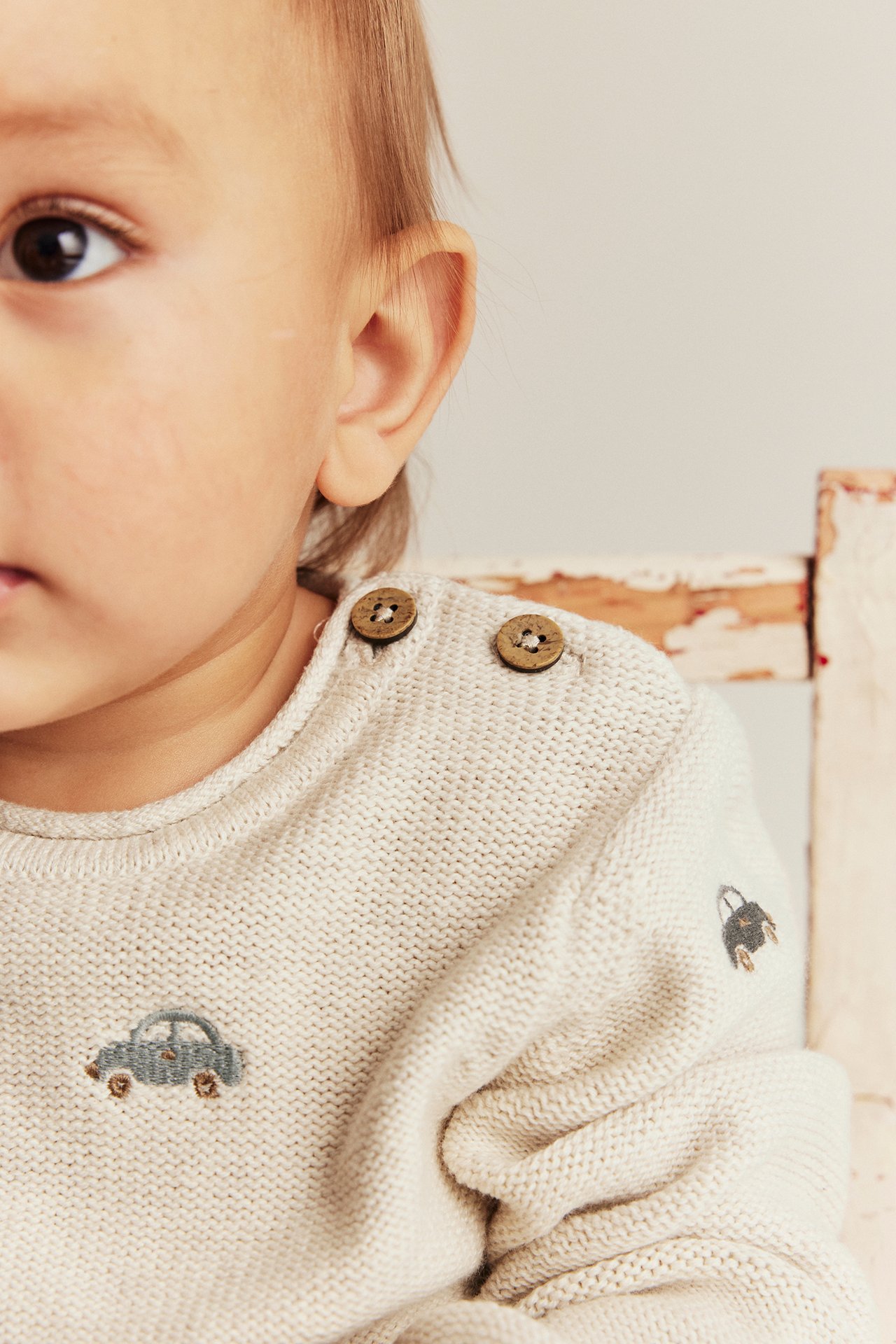 Close-up of baby in beige melange knitted cotton sweater with embroidered cars and wooden shoulder buttons.