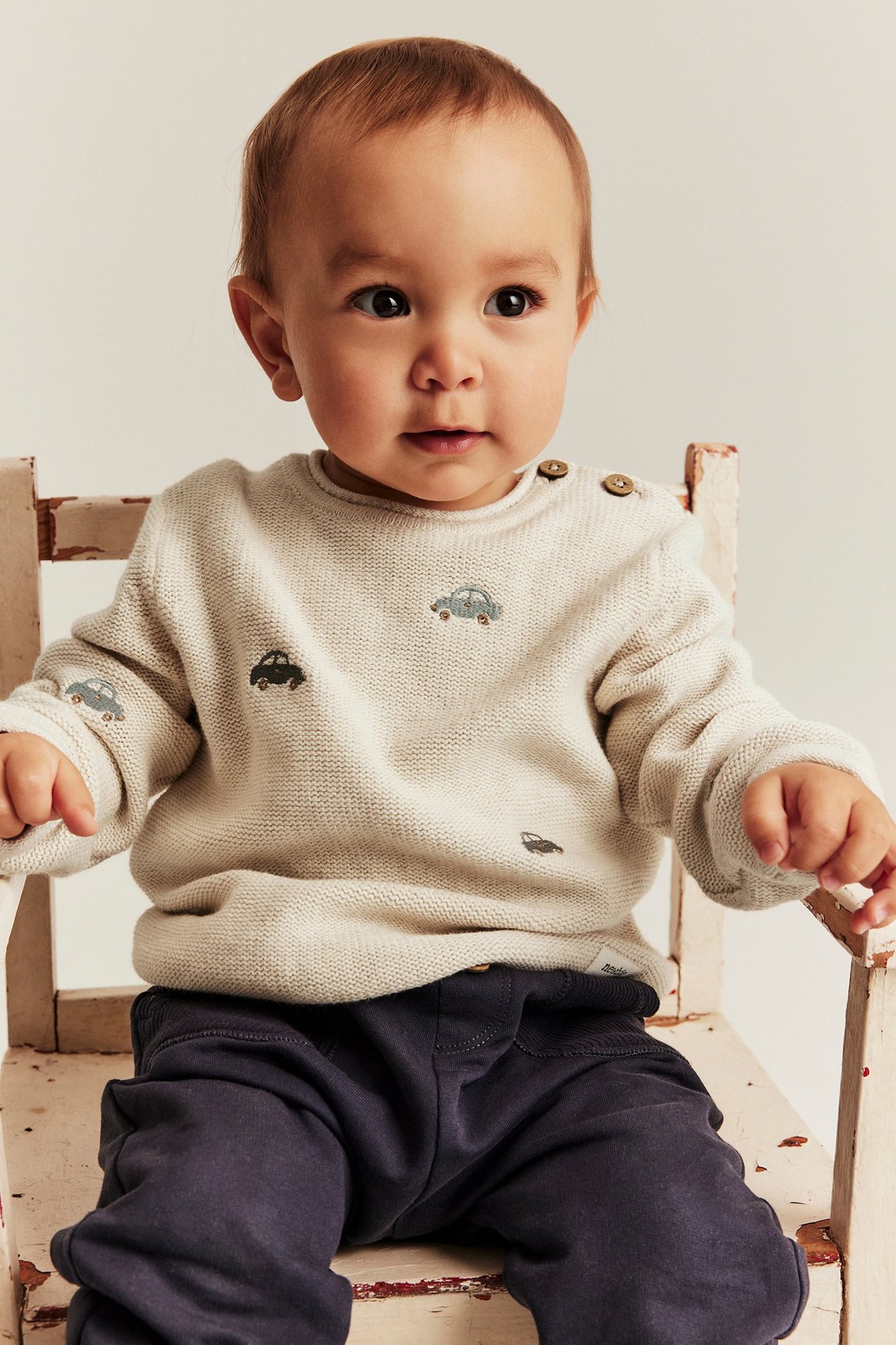 Baby in a beige melange knitted sweater with embroidered cars, shoulder buttons, and dark blue trousers.