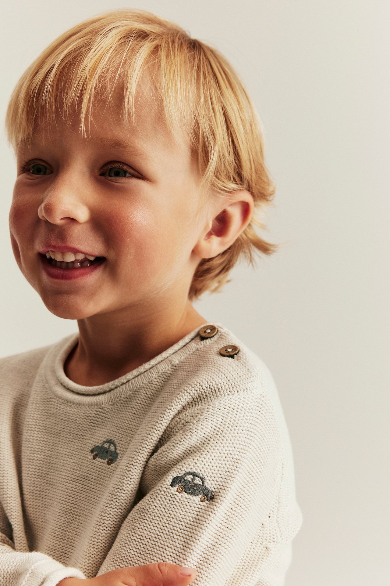 Nahaufnahme eines lächelnden Kindes in einem beigen Strickpullover aus Baumwolle mit Stickerei-Autos und Schulterknöpfen.