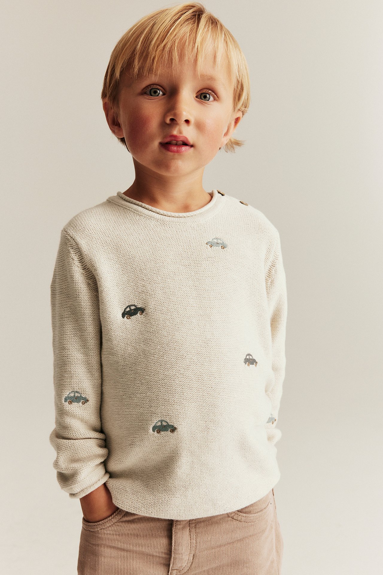 Front view of a kid wearing a beige melange knitted cotton sweater with embroidered cars and brown corduroy pants.