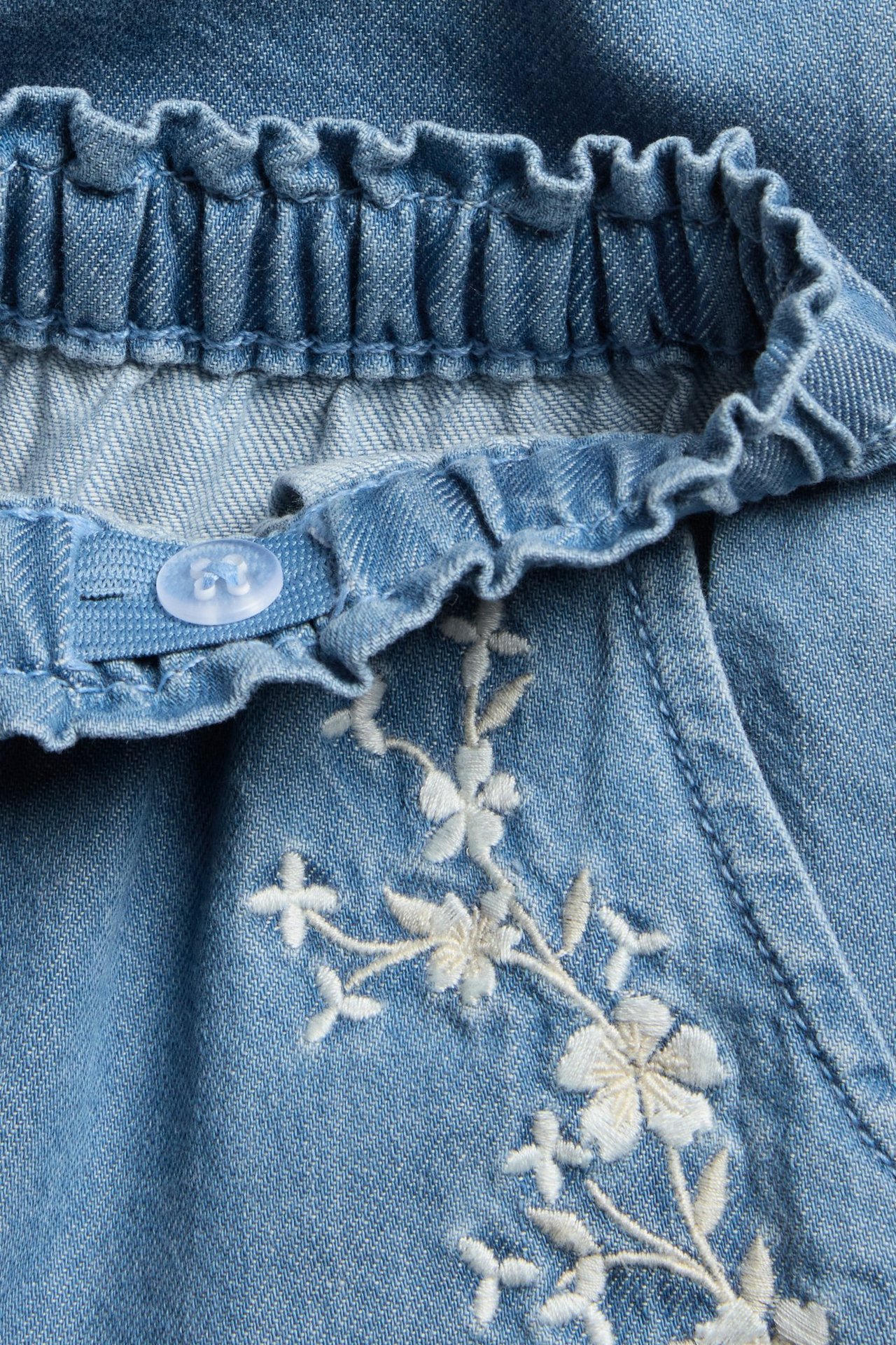 Close-up of kid's light blue denim pants with a ruffled elastic waist, button detail, and white floral embroidery.