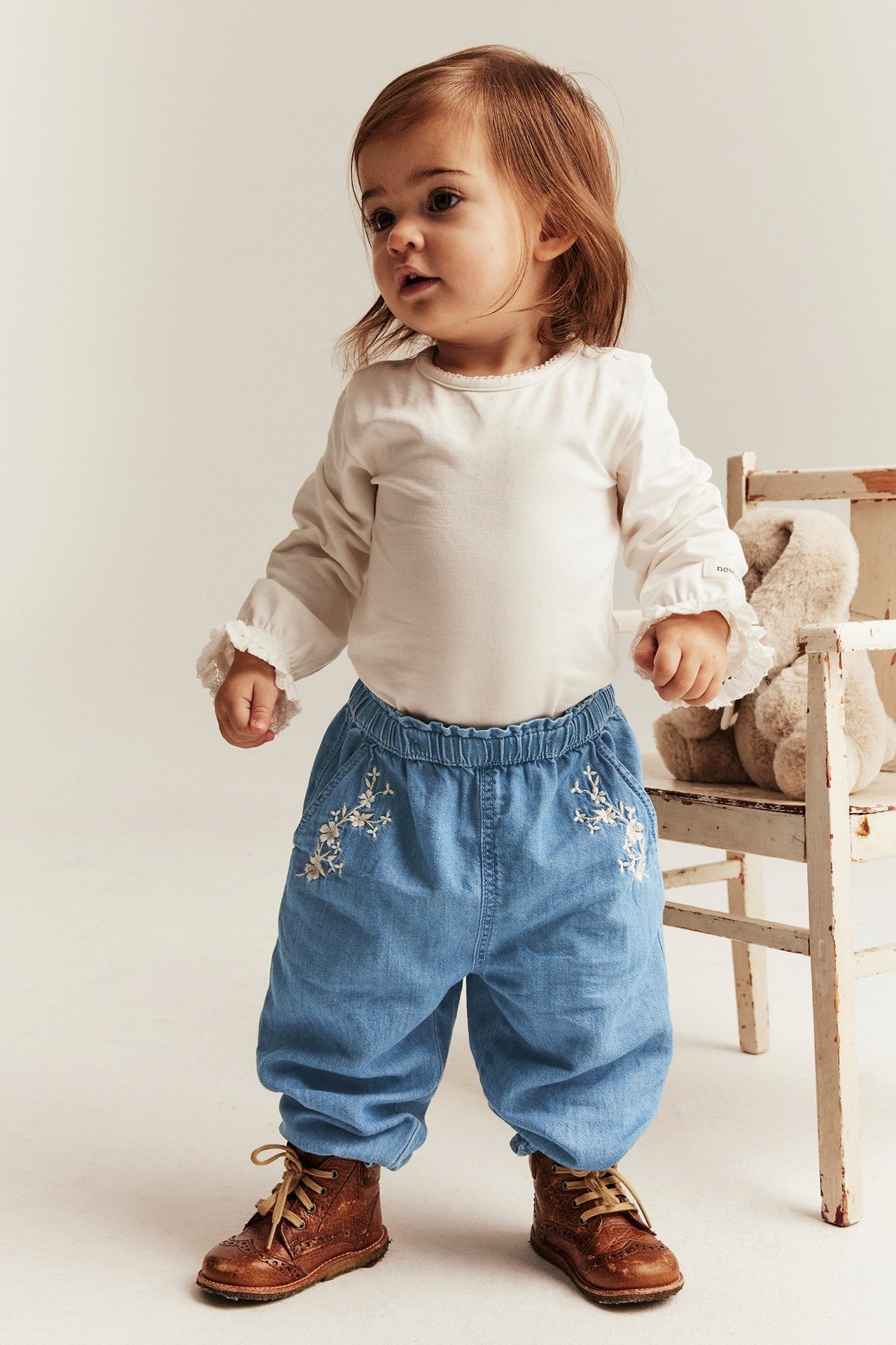 Vorderansicht: Baby in hellblauer Jeanshose mit Blumenstickerei, cremefarbenem Oberteil, braunen Stiefeln.