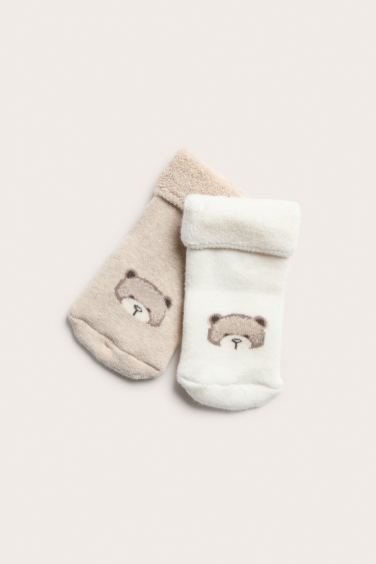 Top view of two baby terry socks, one off-white, one light beige, both with a folded cuff and teddy bear pattern.