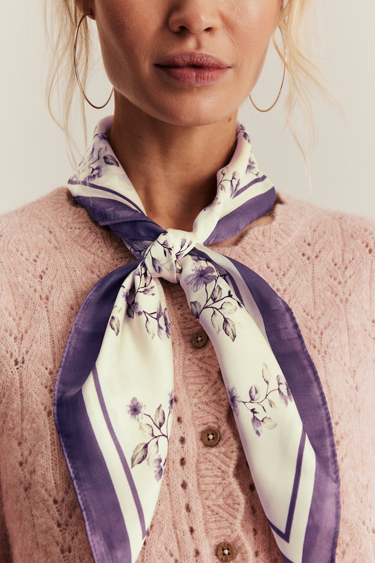 Nahaufnahme: Frau trägt helllila Blumenmuster-Polyesterschal gebunden, goldene Creolen und hellrosa Strickjacke.