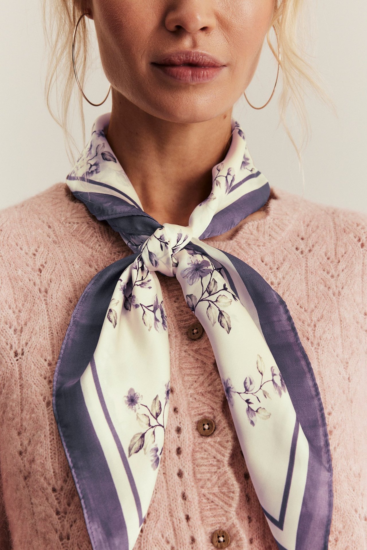 Vorderansicht: Frau trägt helllila Schal mit Blumenmuster aus Polyester, über rosafarbener Strickjacke, dazu goldene Creolen.