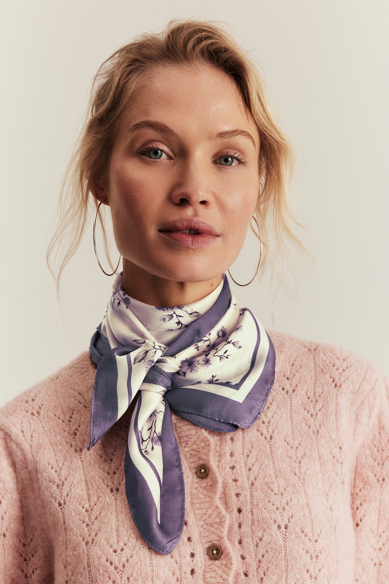 Nahaufnahme einer Frau mit einem weiß-lila geblümten Polyestertuch über einer rosa Strickjacke von vorne.