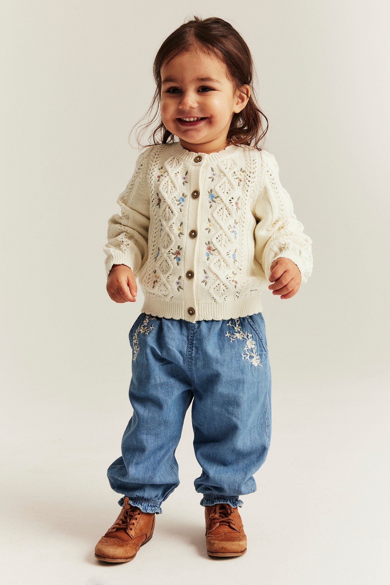 Front view of a smiling baby wearing an off-white cotton floral embroidered cardigan, blue trousers, and brown shoes.
