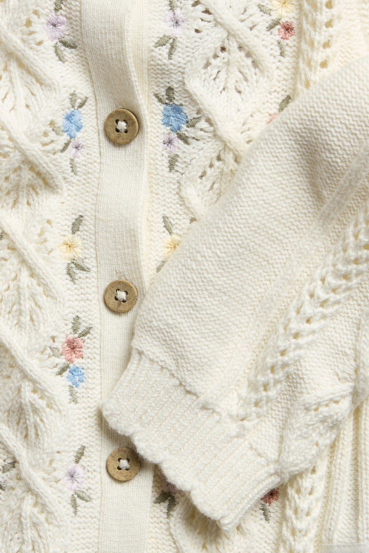 Close-up front view of an off-white cable-knit cotton cardigan with floral embroidery and brown buttons.