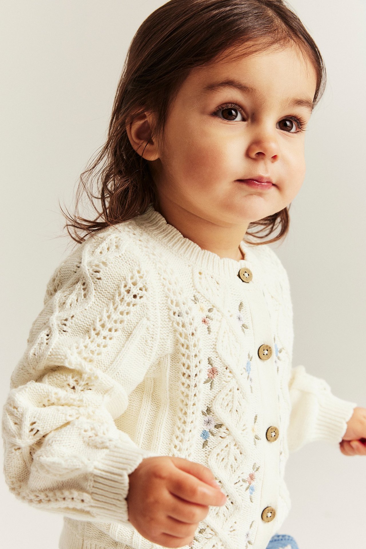 Nahaufnahme, Vorderansicht, eines Babys in einer offwhite gemusterten Strickjacke aus Baumwolle mit Blumenstickerei und Holzknöpfen.