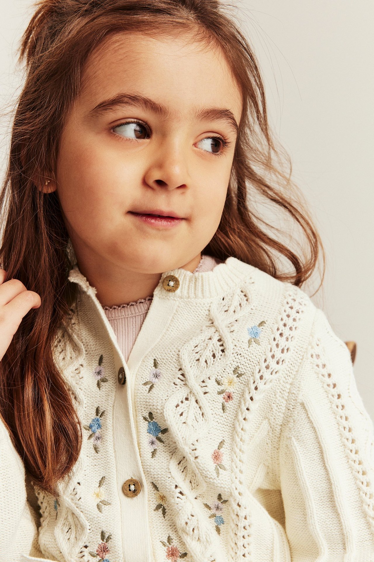 Close-up of a kid wearing an off-white floral embroidered cotton cardigan with buttons over a pink top.
