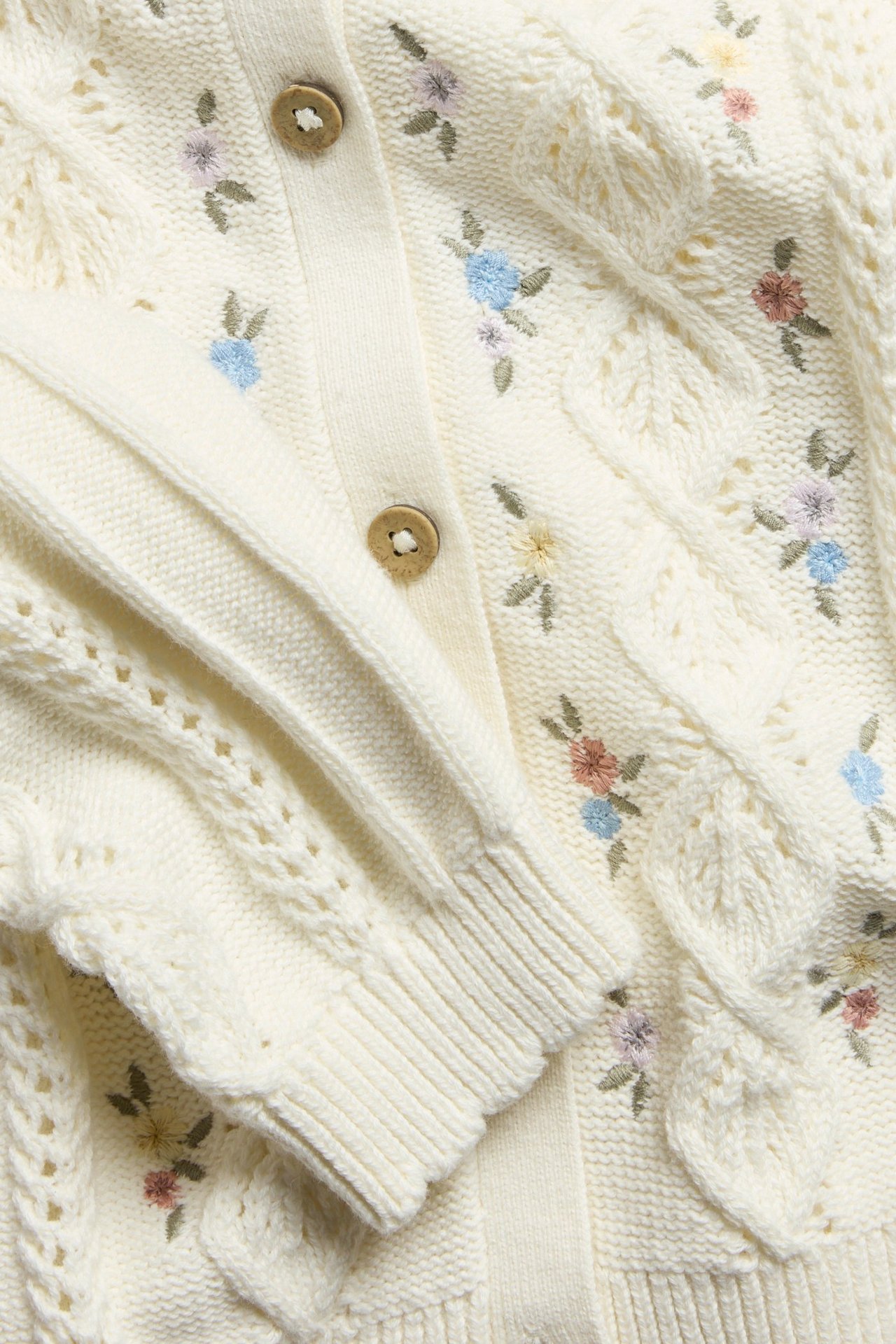 Close-up of an off-white cotton cable-knit cardigan with colorful floral embroidery and wooden buttons. Ribbed cuffs.