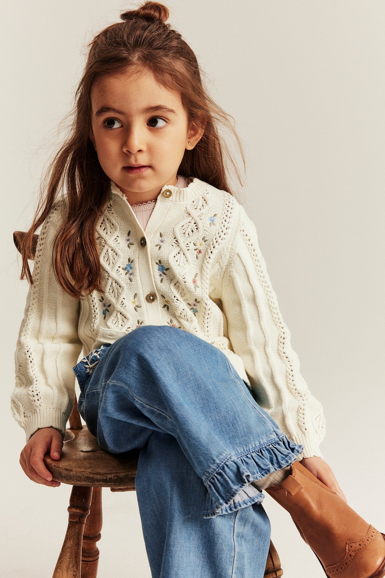 Front view: Kid in an off-white embroidered cotton cardigan with buttons, blue jeans, and brown boots.