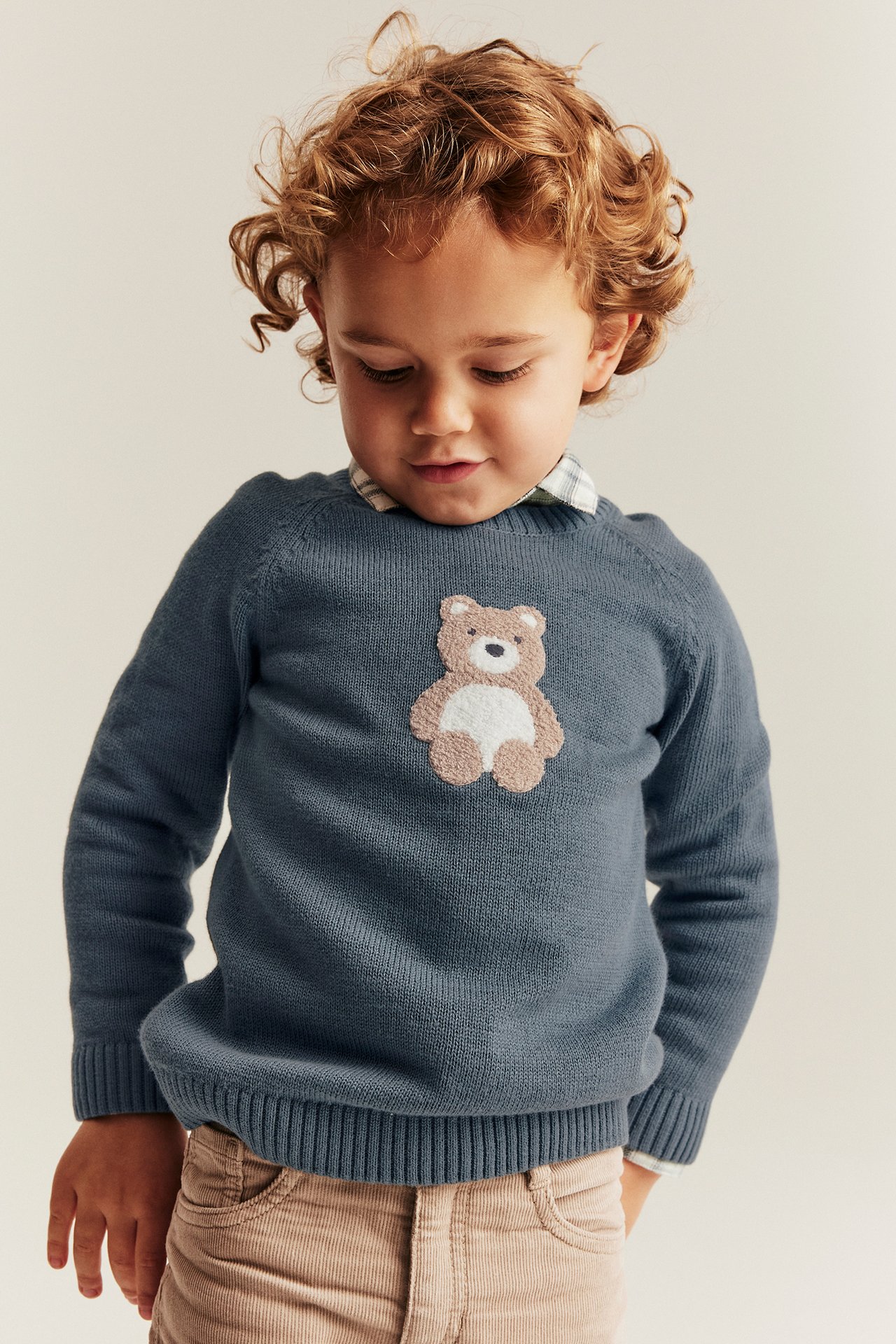 Front view of a kid in a blue cotton sweater with a bear application, worn over a plaid shirt.