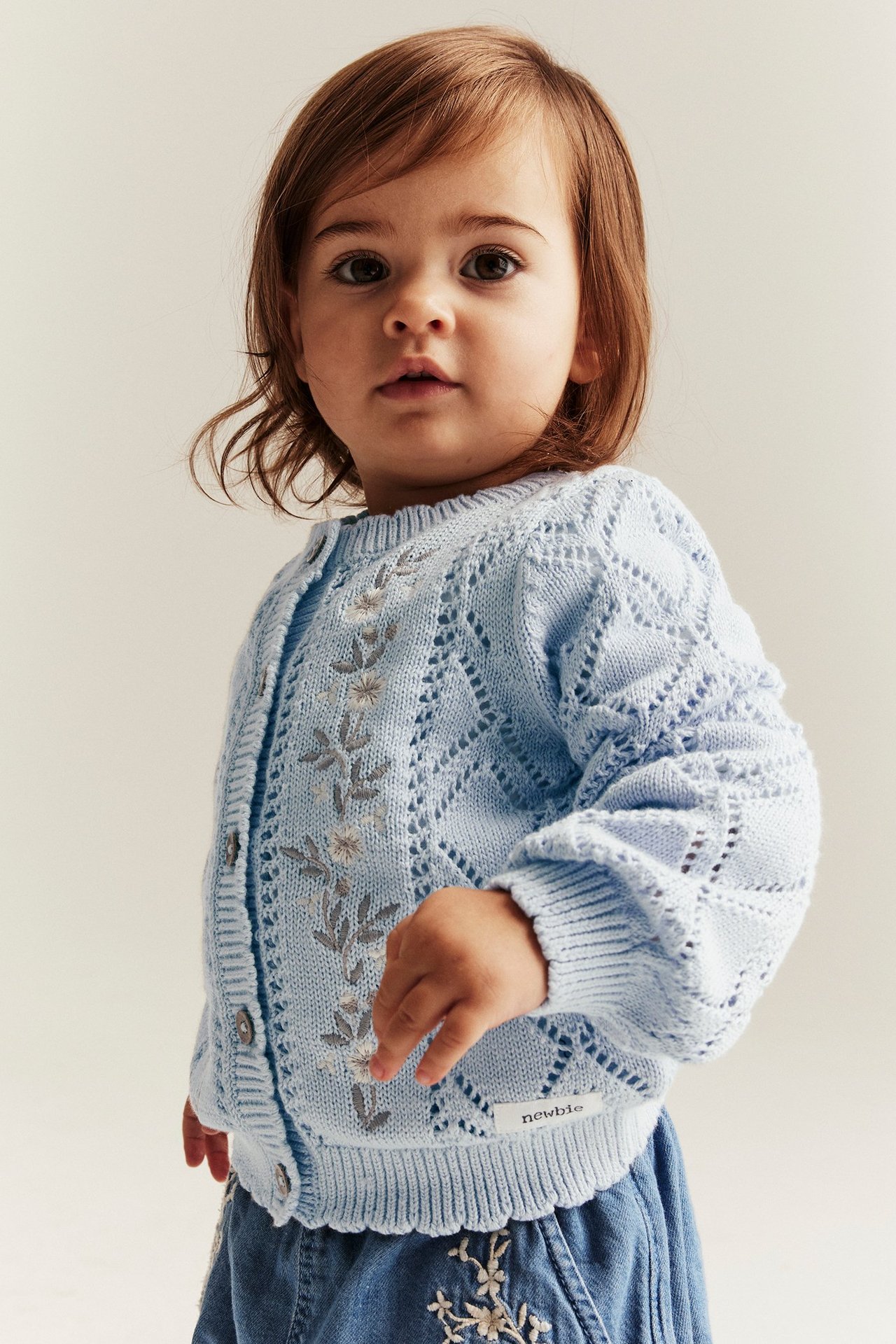 Vorderansicht eines Babys in hellblauer gestrickter Baumwollstrickjacke mit Blumenstickerei und blauem Rock.
