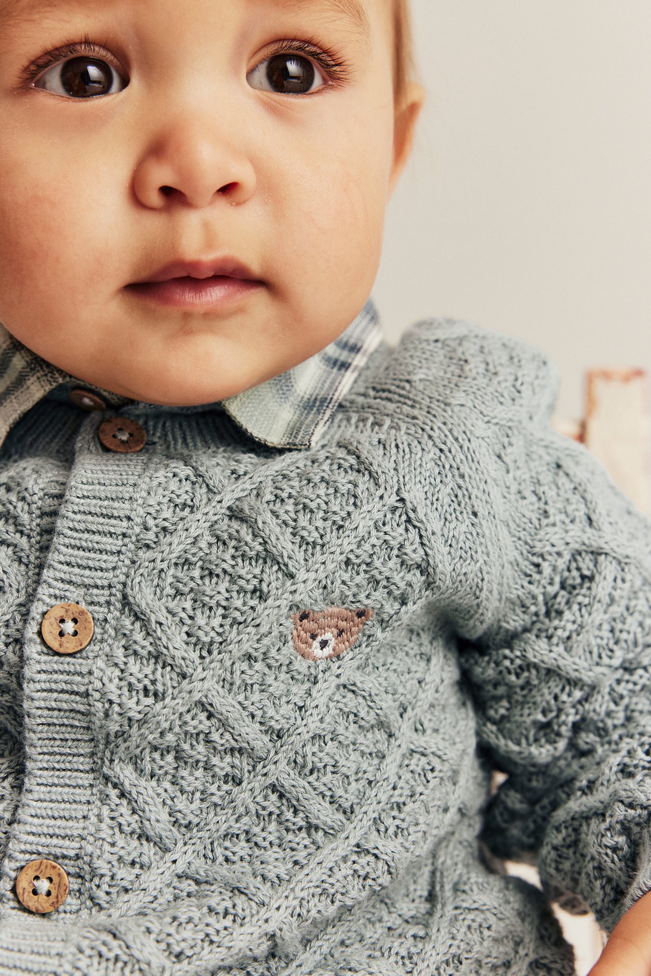 Close-up of a baby wearing a light green knitted cardigan with a teddy bear embroidery over a plaid shirt.