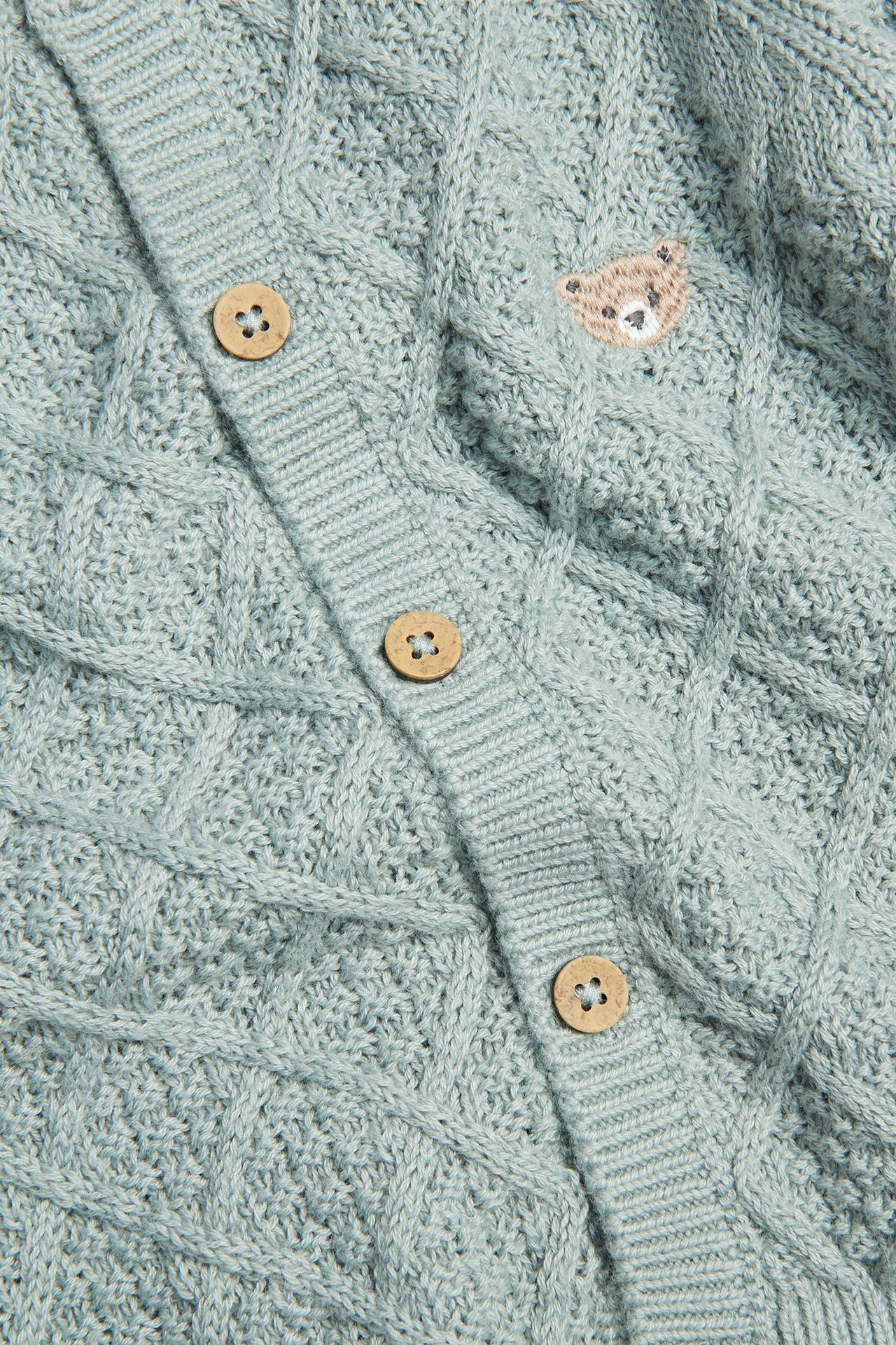 Close-up of a baby's light green cotton cable knit cardigan with wooden buttons and a teddy bear embroidery.