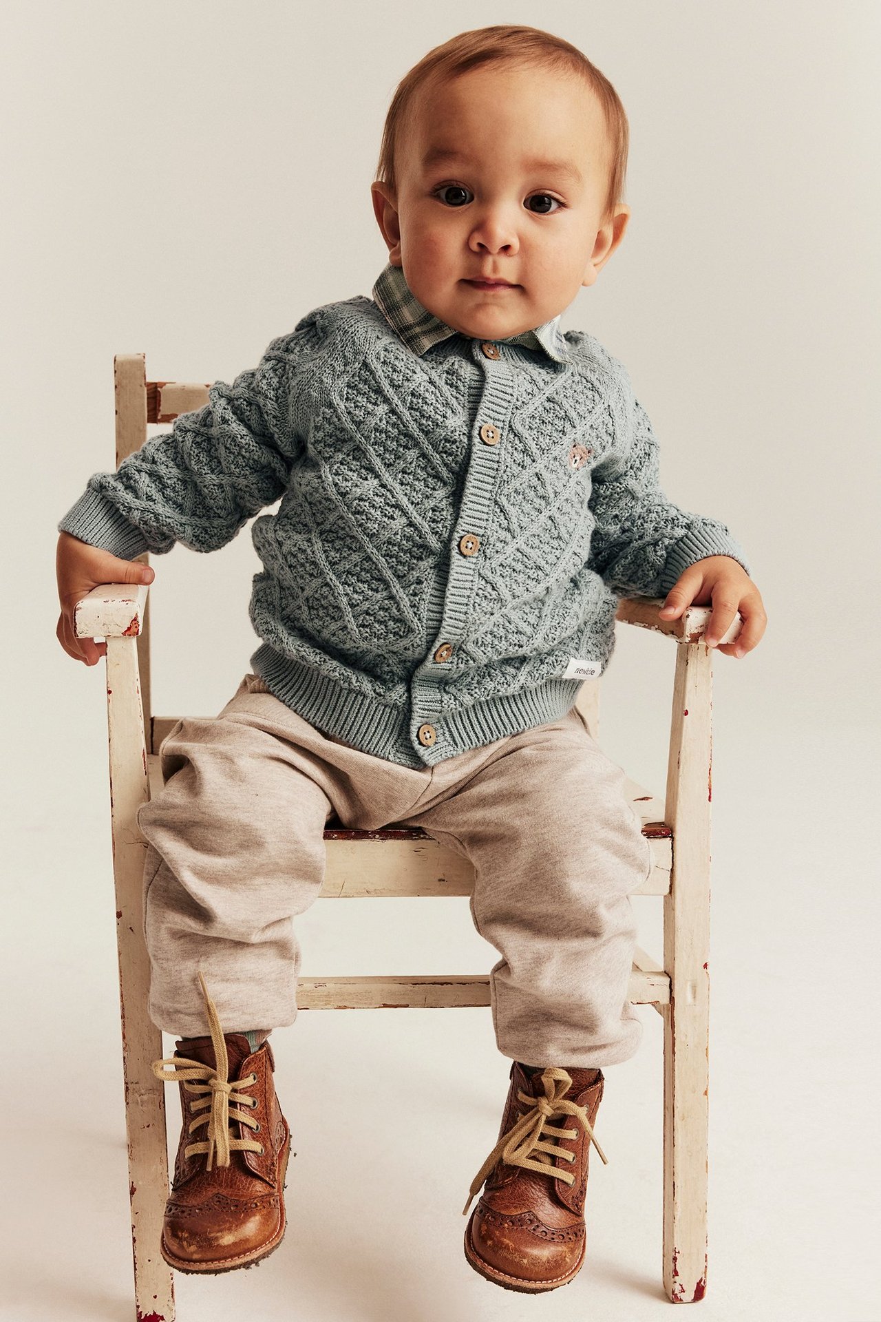 Vorderansicht: Baby trägt hellgrüne gestrickte Rautenmuster-Strickjacke mit Holzknöpfen, kariertes Hemd, beige Hose, braune Stiefel.