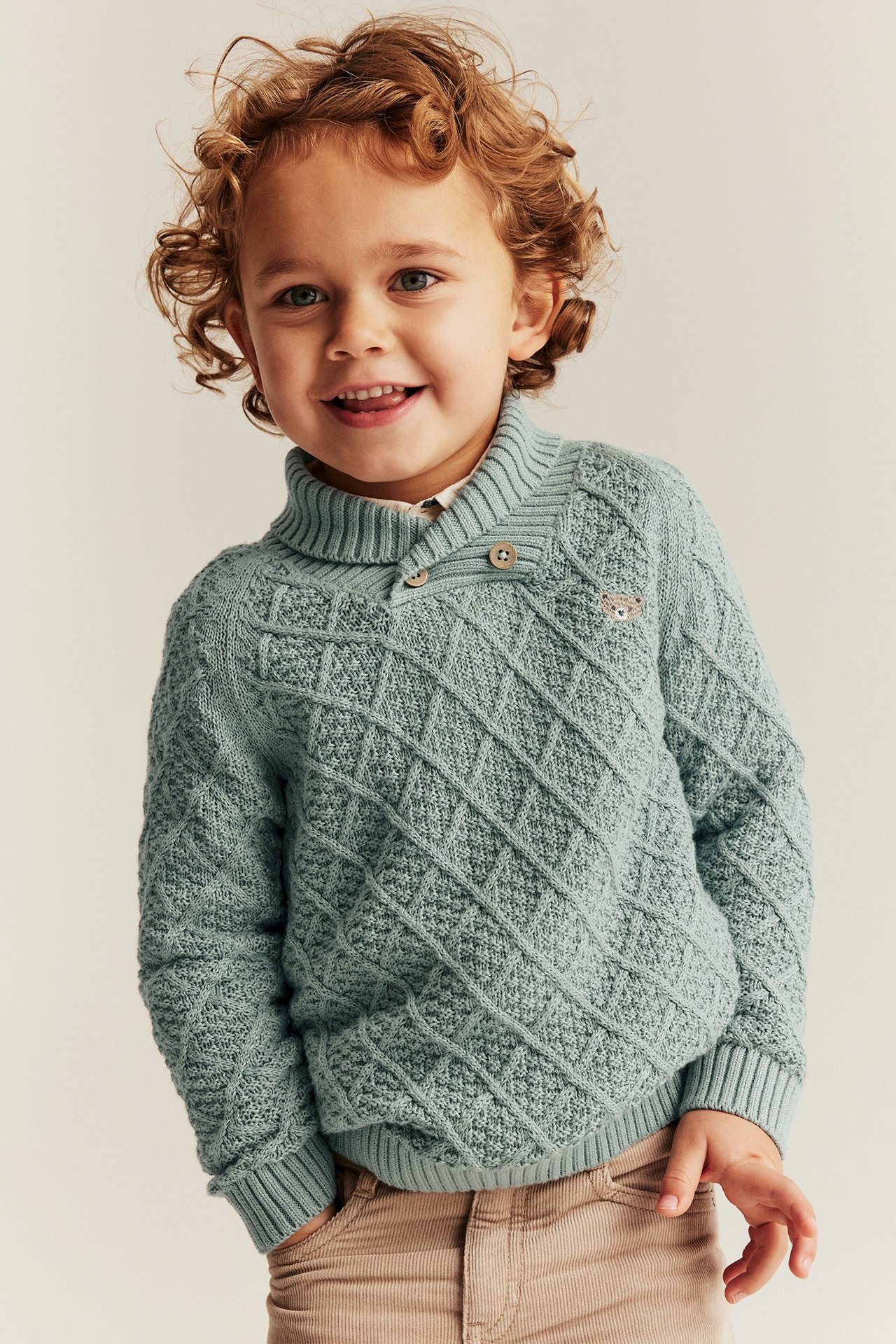 Vorderansicht eines Kindes in hellgrünem Strickpullover mit Knöpfkragen, Teddy-Stickerei und beiger Hose.