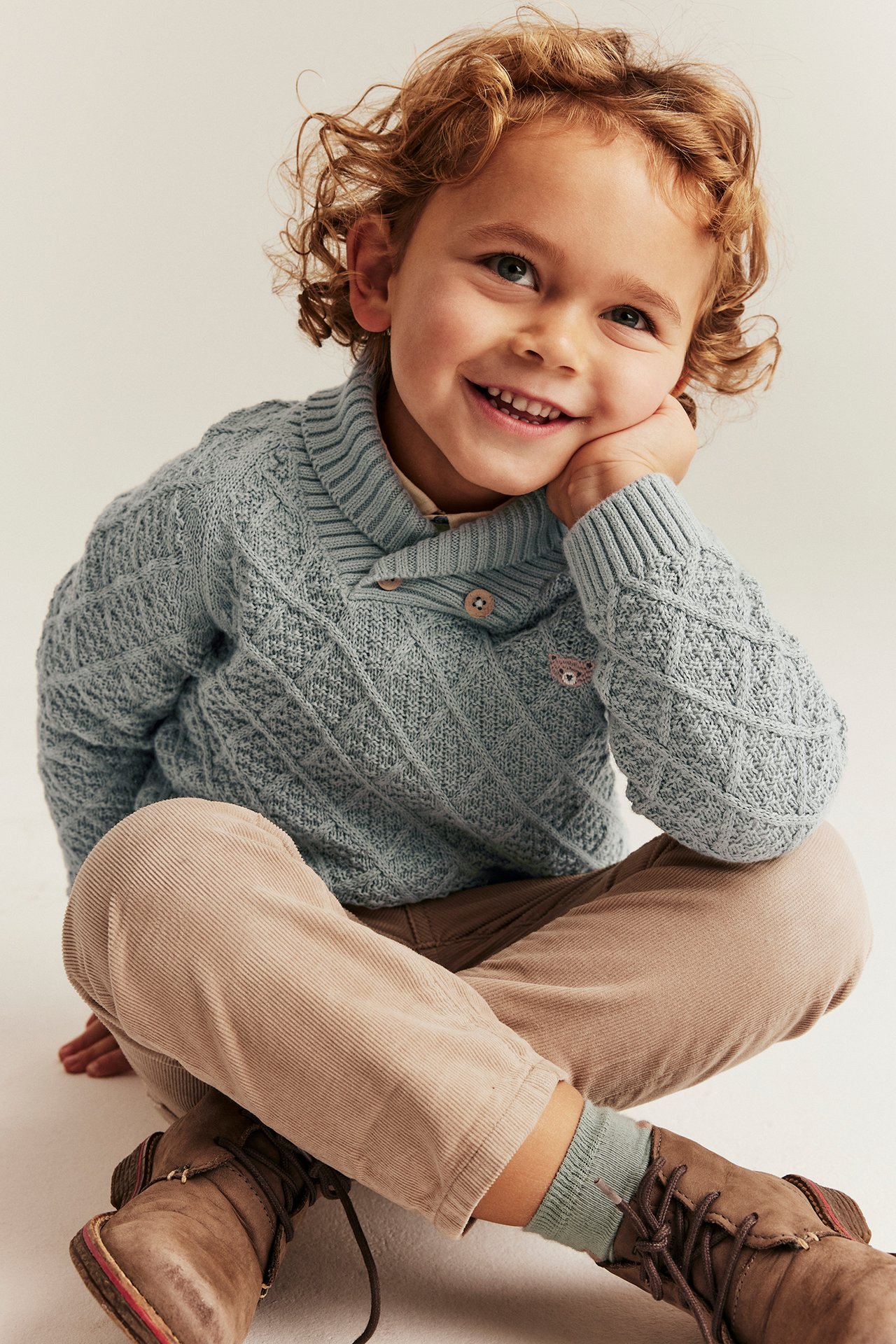 Vorderansicht: Kind in hellgrünem gestricktem Baumwollpullover mit Teddy-Applikation, Schalkragen, beiger Cordhose, braunen Stiefeln.