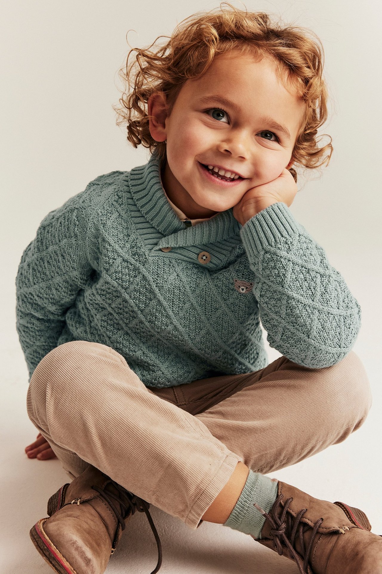 Vorderansicht eines Kindes in hellgrünem Strickpullover mit Bärenapplikation, beiger Cordhose und braunen Stiefeln.