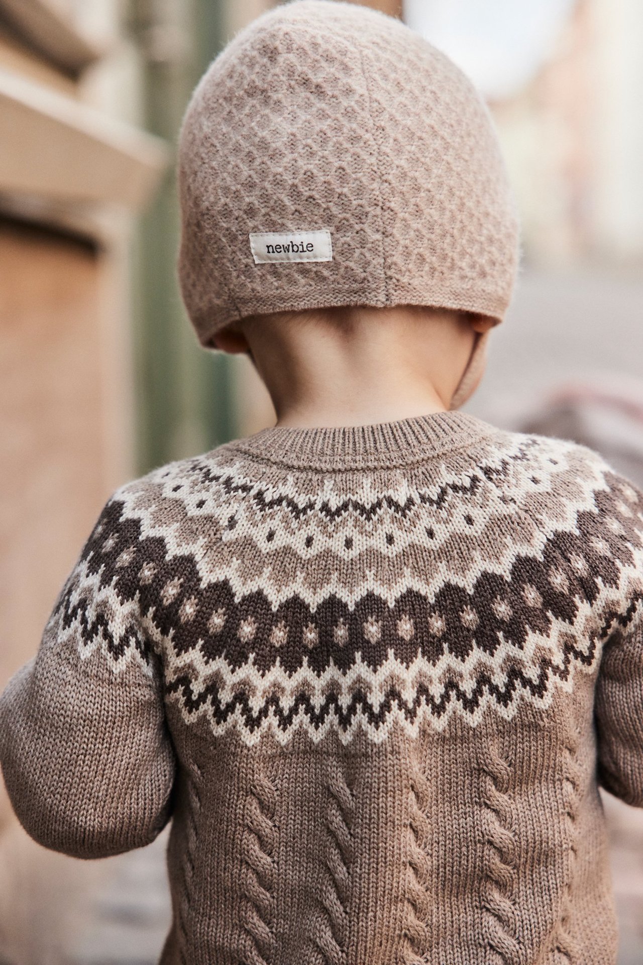Rückenansicht eines Babys in beigefarbener gemusterter Strick-Helmmütze und Fair-Isle-Pullover.
