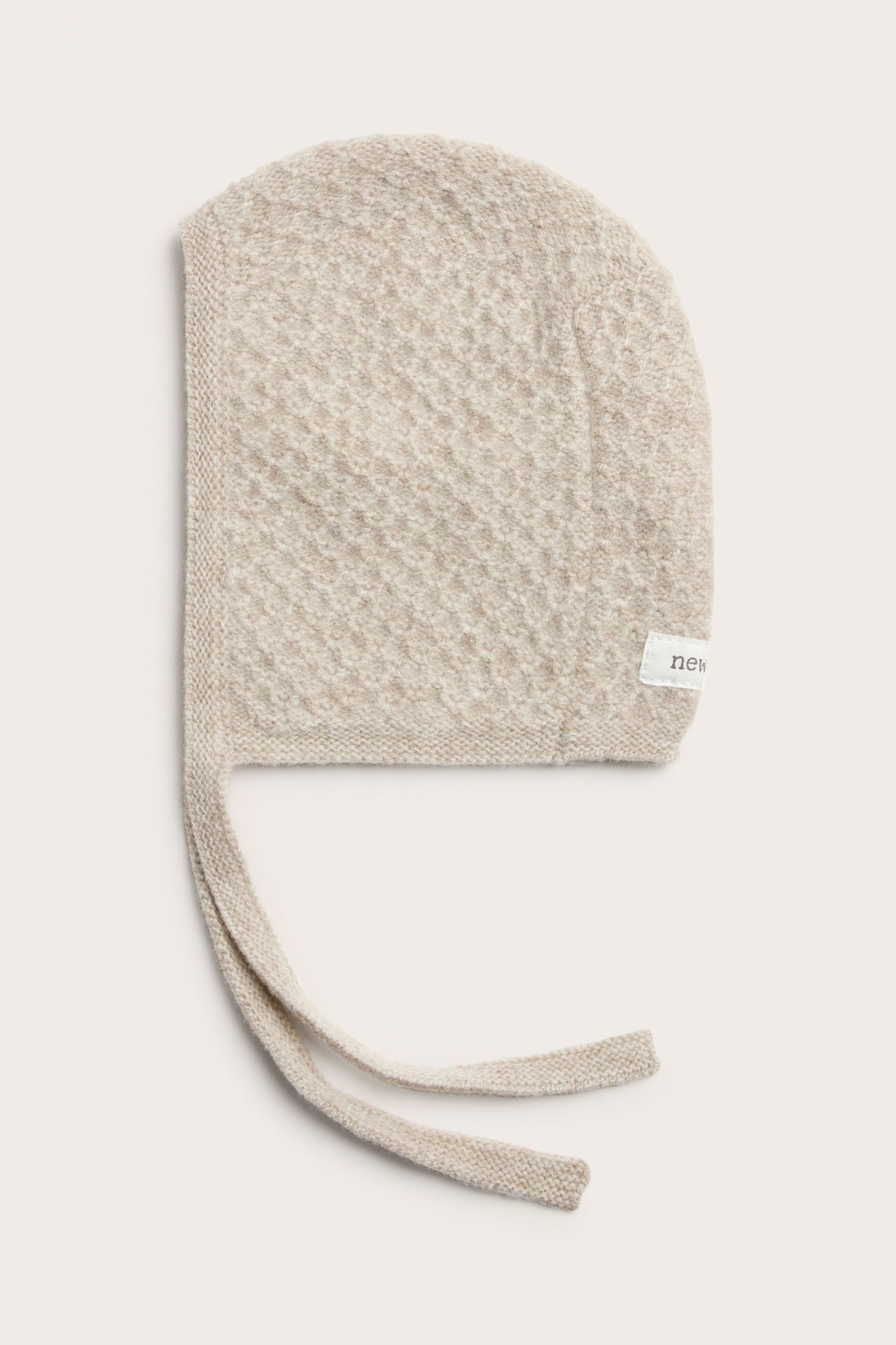 Flach liegende hellbeige strukturierte Strick-Babyhaube mit Bändern.