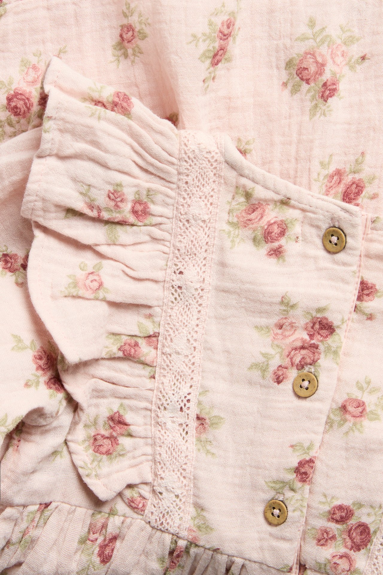 Close-up of a light pink floral muslin dress with ruffles, lace trim, and visible wooden buttons.
