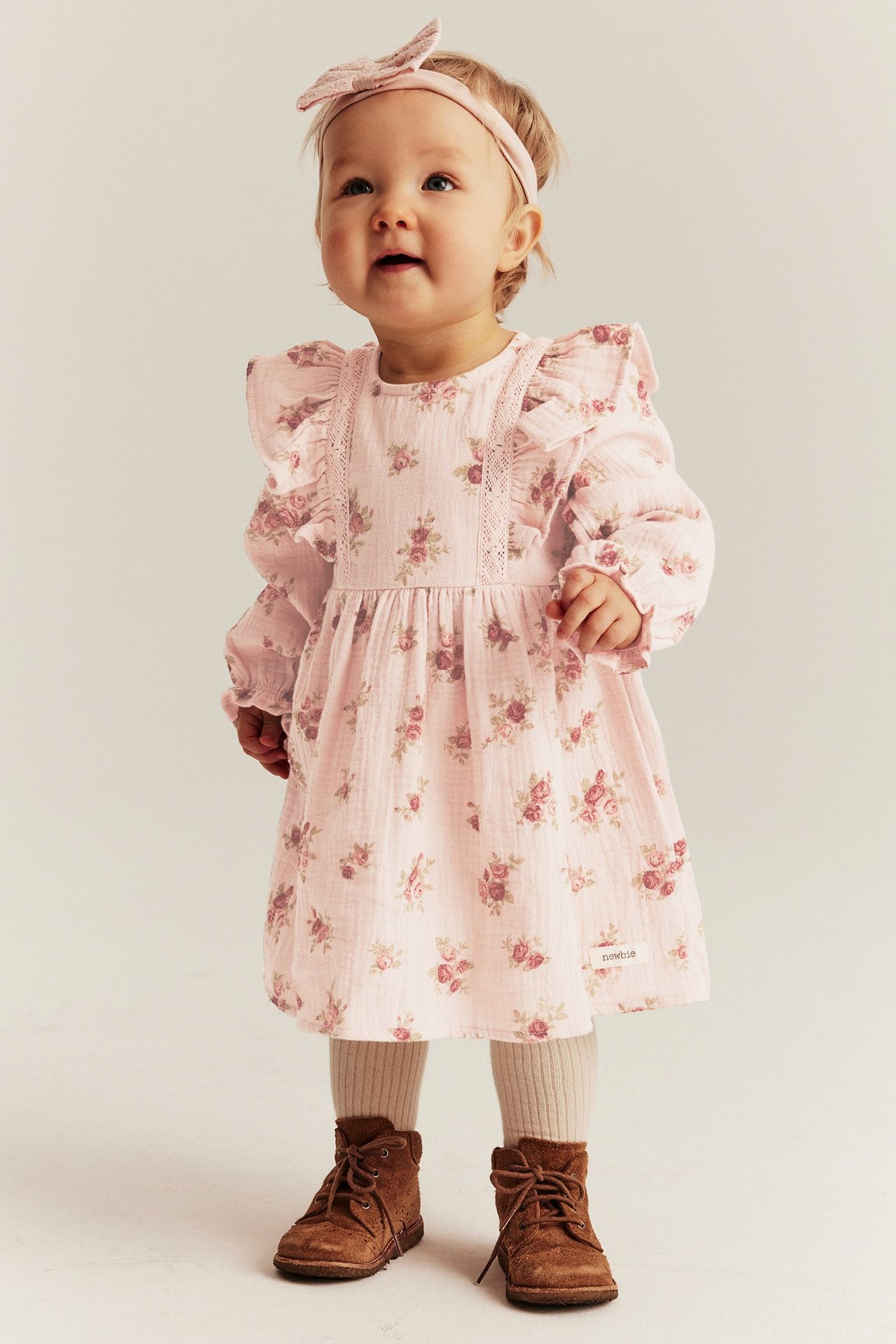 Baby wears a light pink floral muslin dress with ruffles, matching bow headband, beige tights, and brown boots.