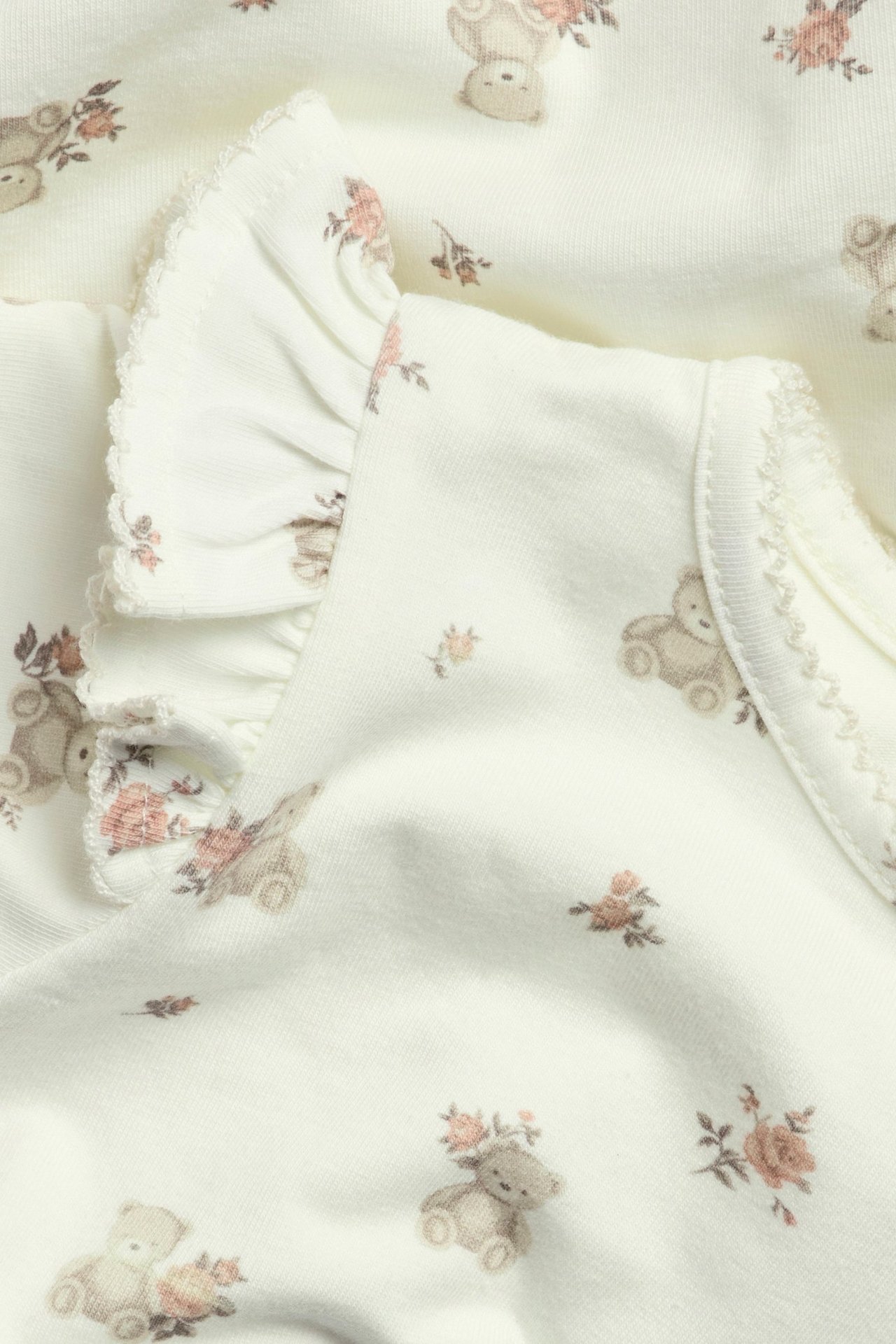 Nahaufnahme: Off-white Baby-Jersey-Body mit Bären- und Blumenmuster, Rüschen und Picotbesatz.