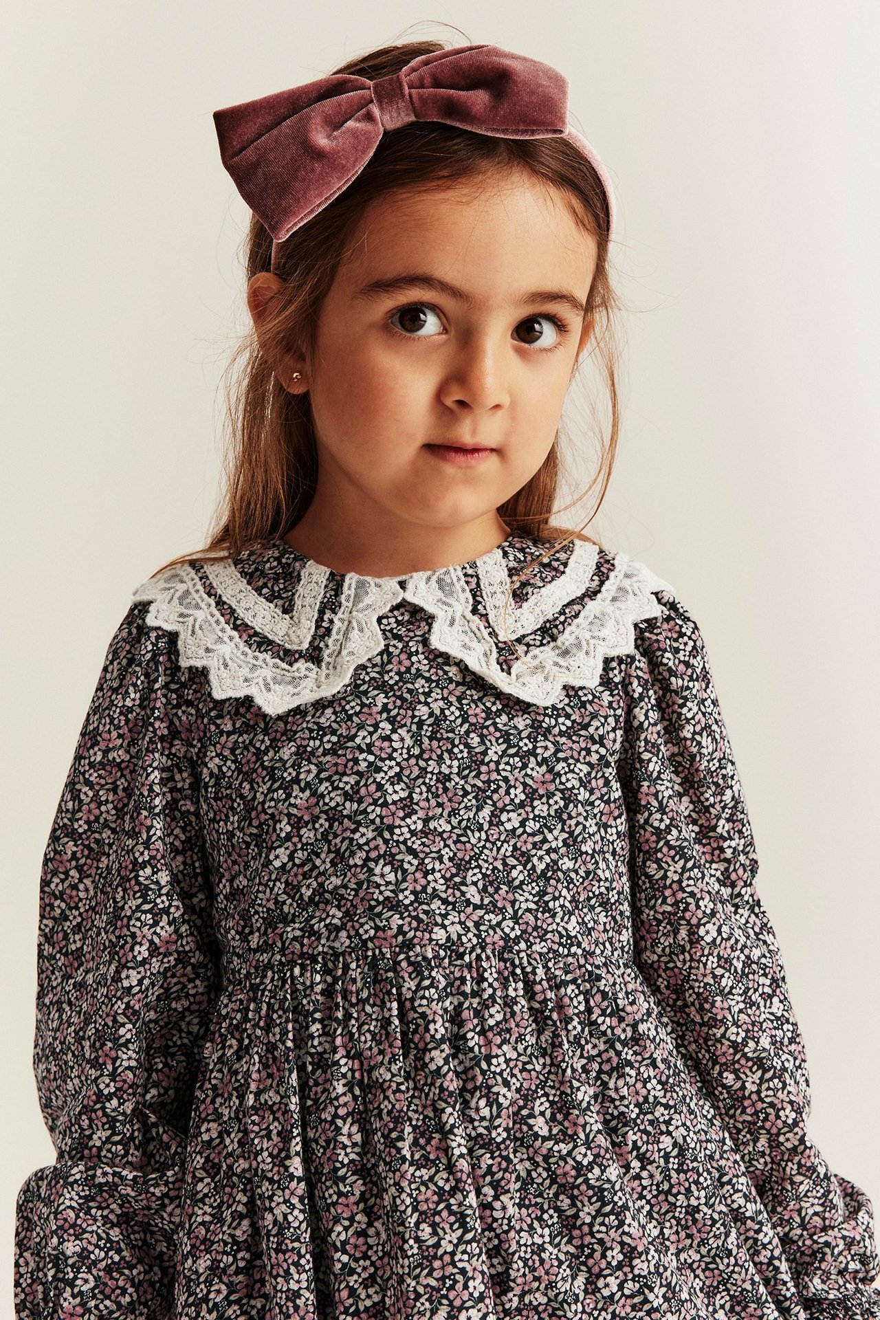Close-up of a kid wearing a black floral dress with a white lace collar and a pink velvet headband.